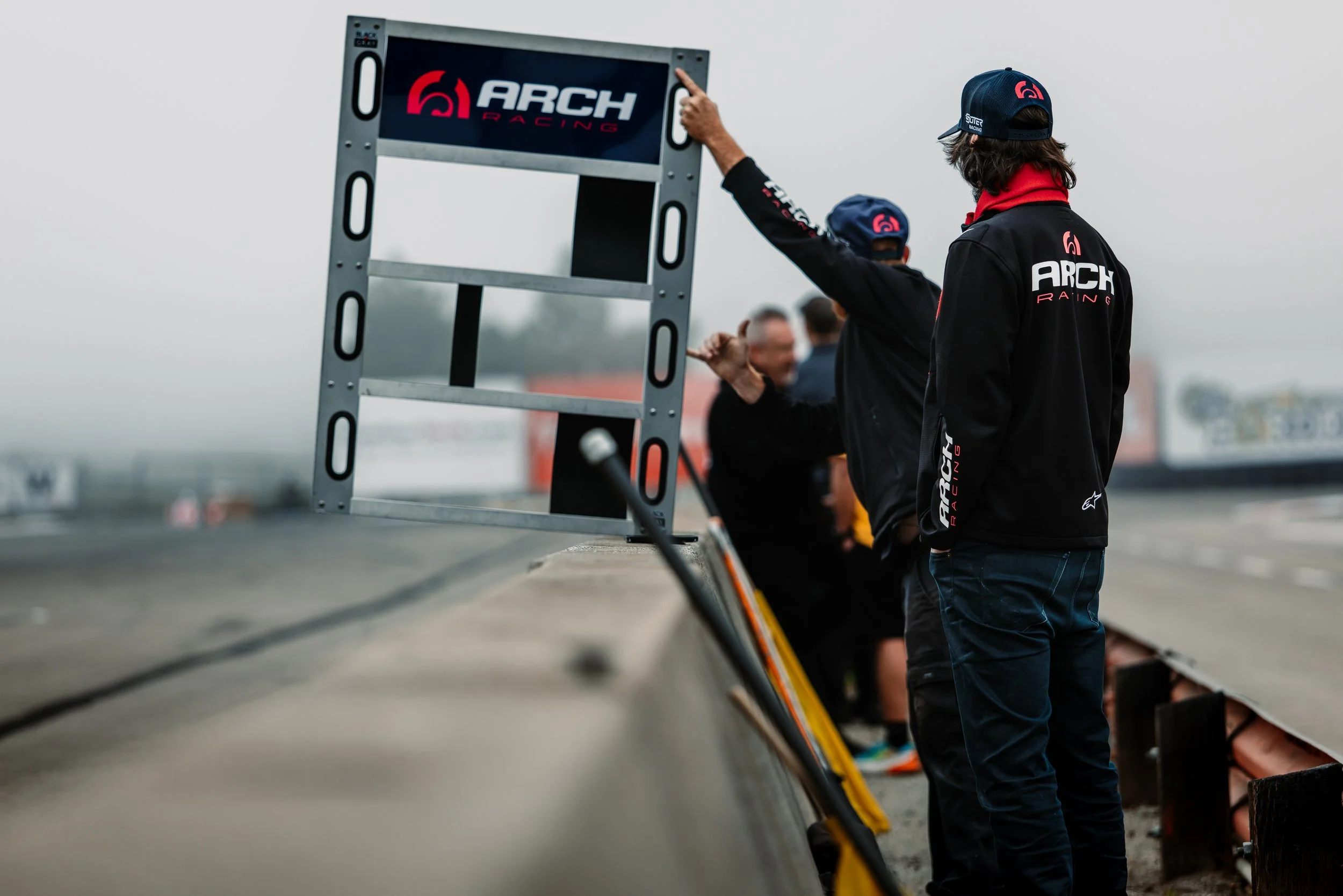 Two men wearing black jackets with 'Arch Racing' logos and caps stand beside a race track, looking at a signboard with 'ARCH RACING' logo, with three blurred people in the background on a cloudy day.