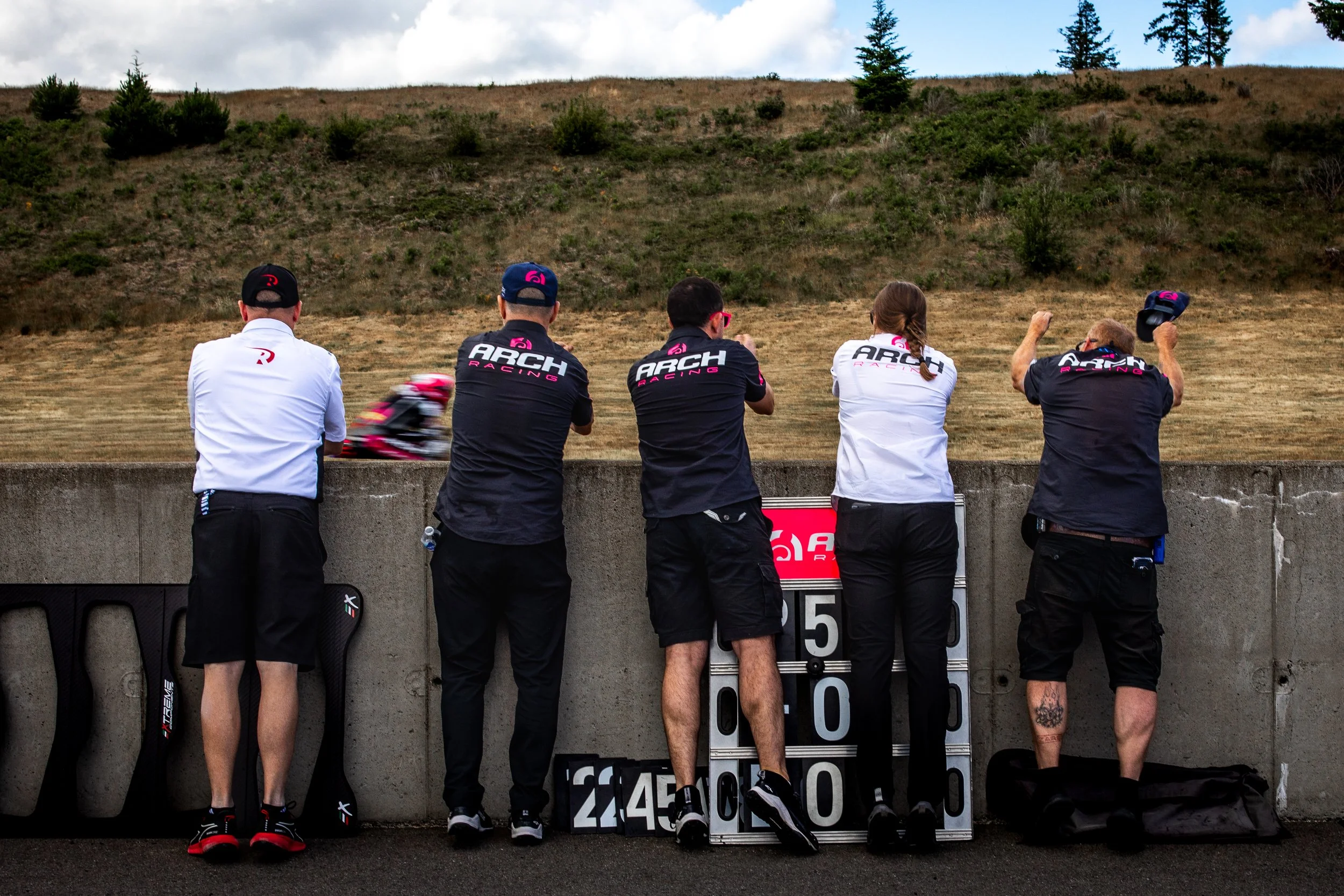 Five race team members, dressed in black and white ARCH RACING uniforms, stand at a concrete barrier watching a blurry red and black motorcycle go by on a racetrack. One person is adjusting his cap, and another is taking a photo with a phone. A score board with numbers is in front of them.
