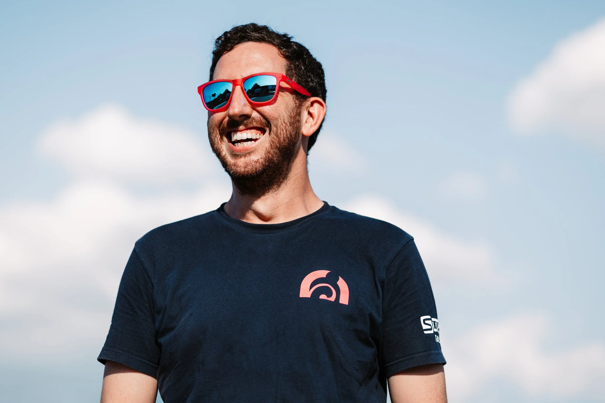 A smiling man with dark curly hair, a beard, and red sunglasses, wearing a black t-shirt, standing outdoors against a partly cloudy sky.