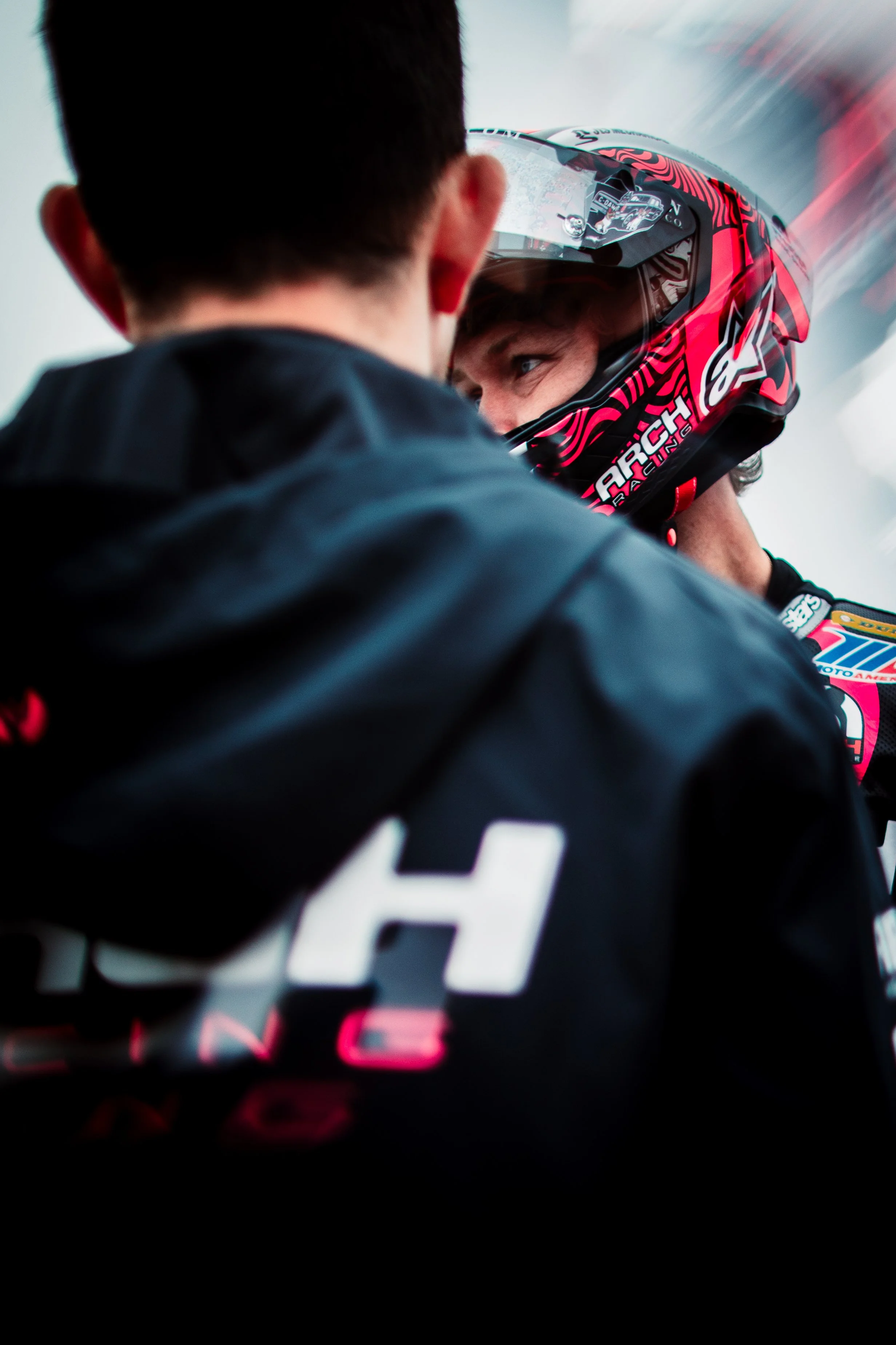A person in racing gear, wearing a red helmet, is looking at a team member who is helping him prepare for a race.
