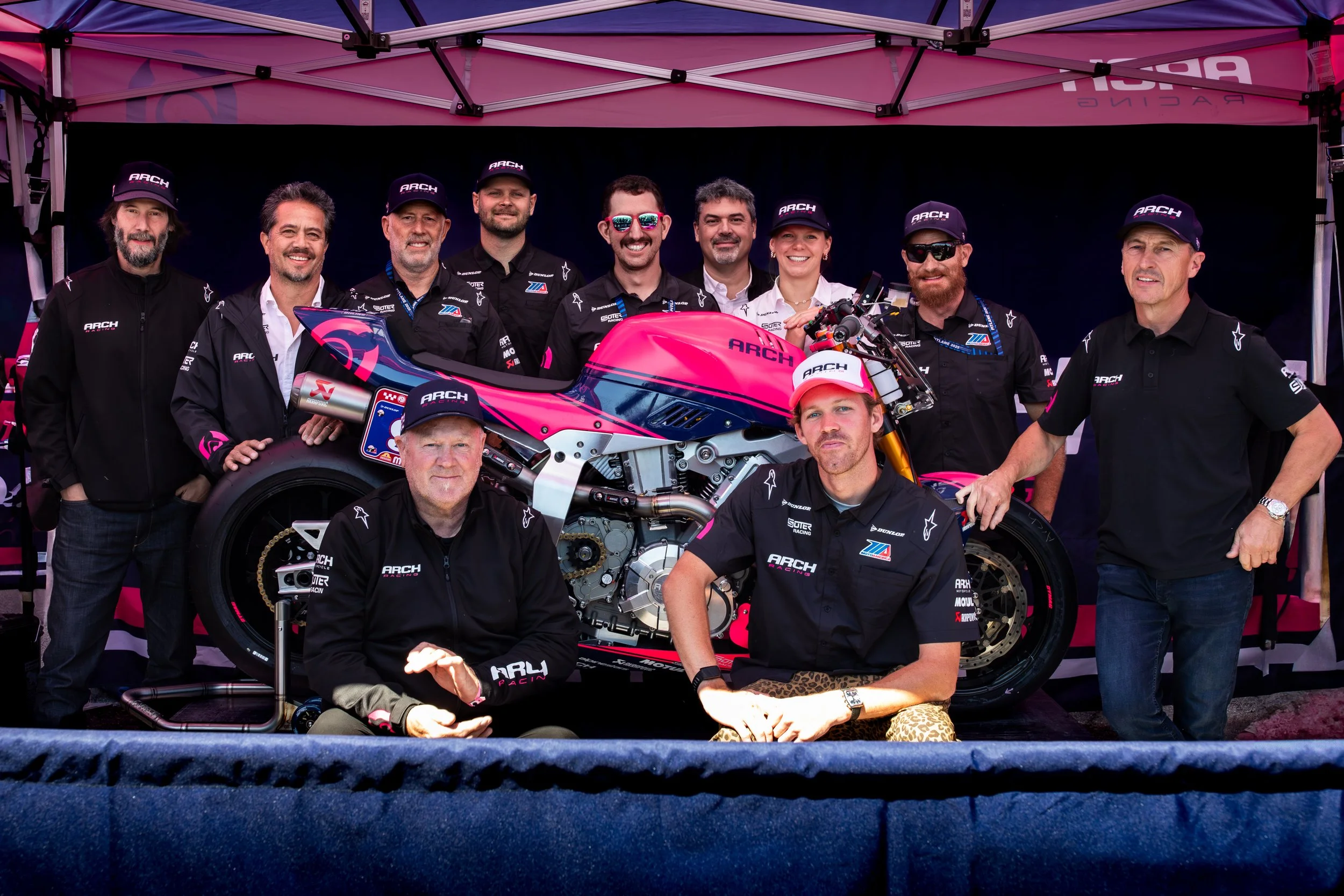 Group of race car team members posing with a pink and blue race motorcycle under a pink canopy, some wearing black team uniforms and hats, others in casual clothes, smiling for the camera.