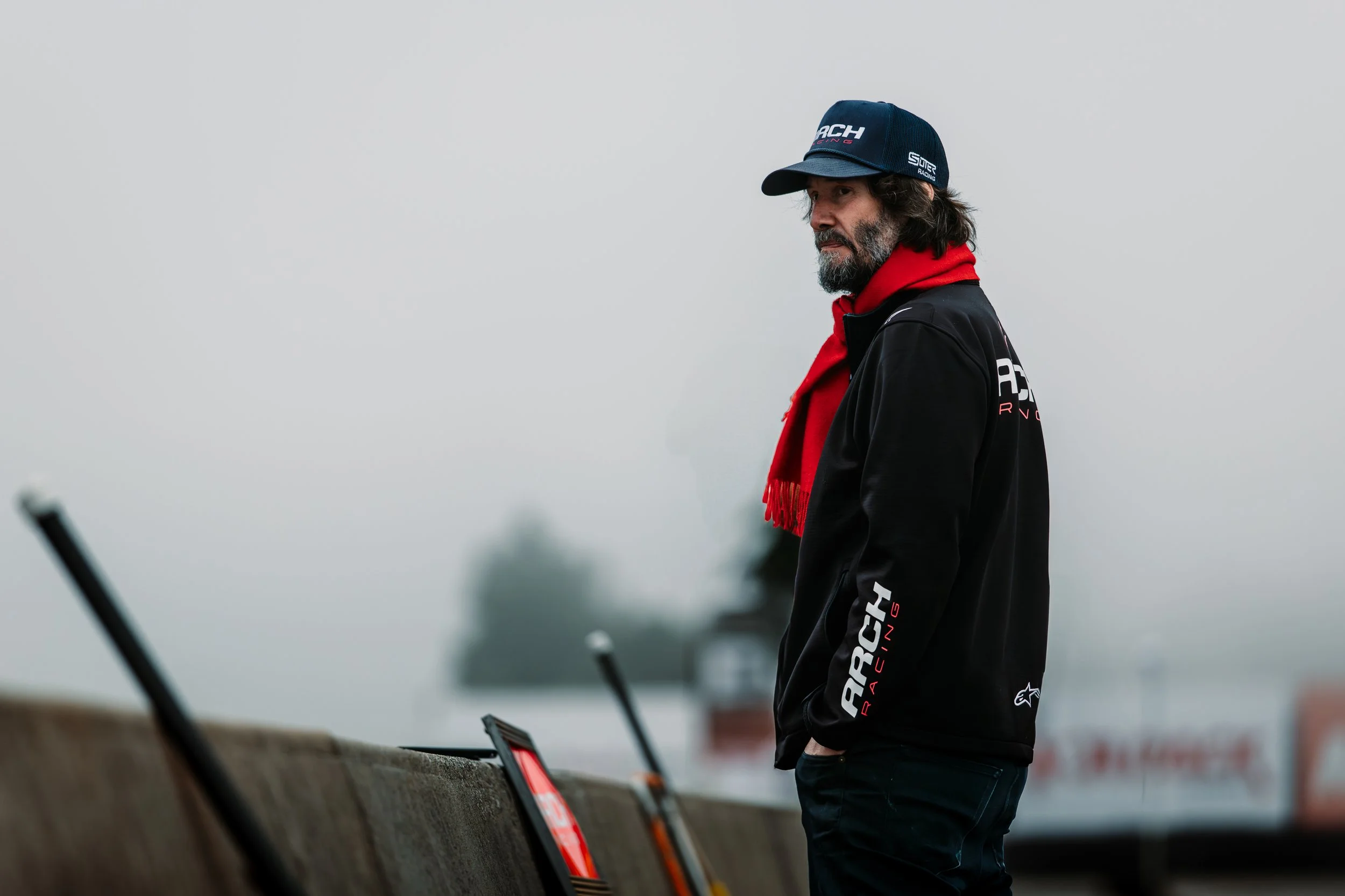 Man in black racing jacket, red hoodie, and blue cap standing by a racetrack on a foggy day.