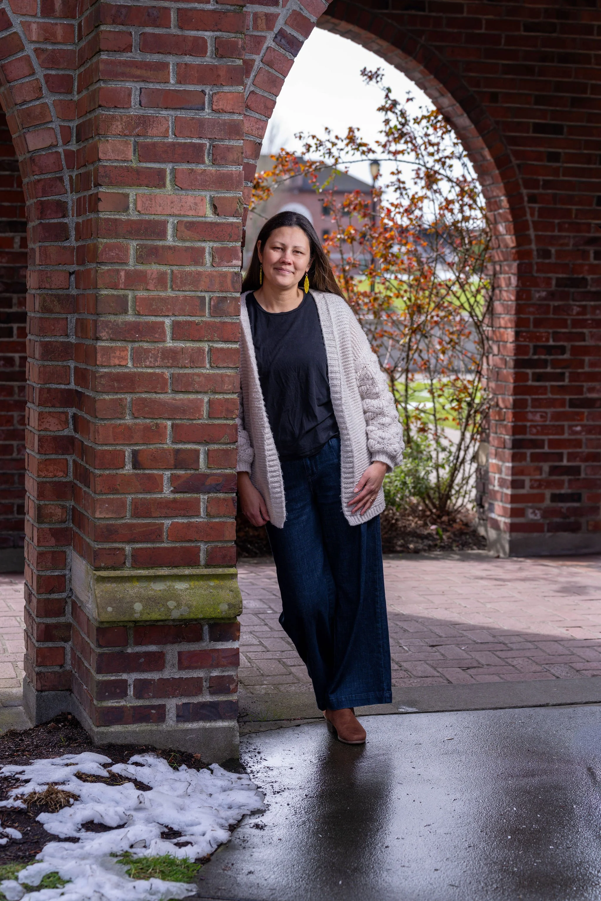 Photo of Hannah smiling and leaning against a brick wall under an arch outside a building.