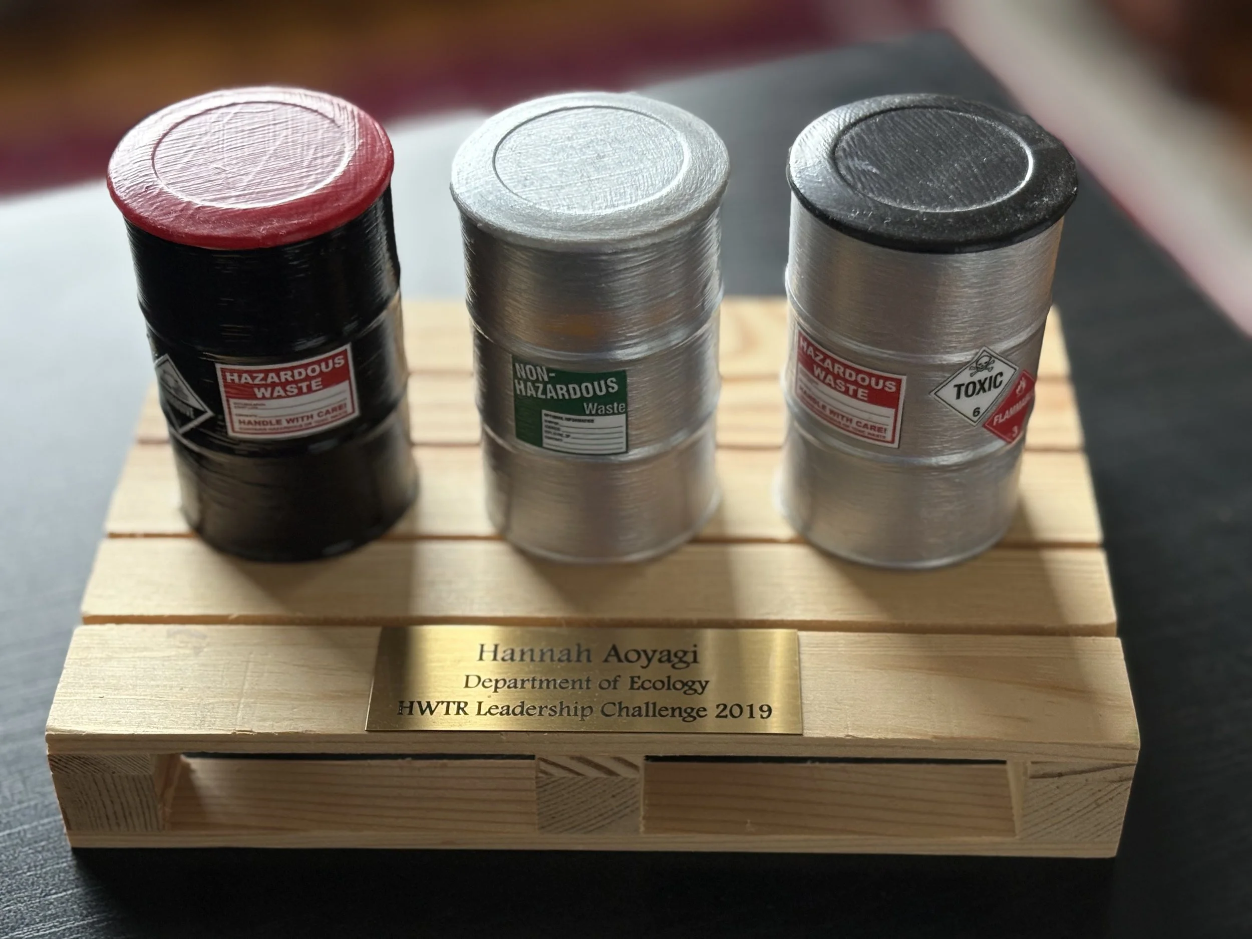 A tiny wooden pallet and three miniature drums of toxic waste with a plaque saying Hannah Aoyagi, Department of Ecology, HWTR Leadership Challenge 2019.