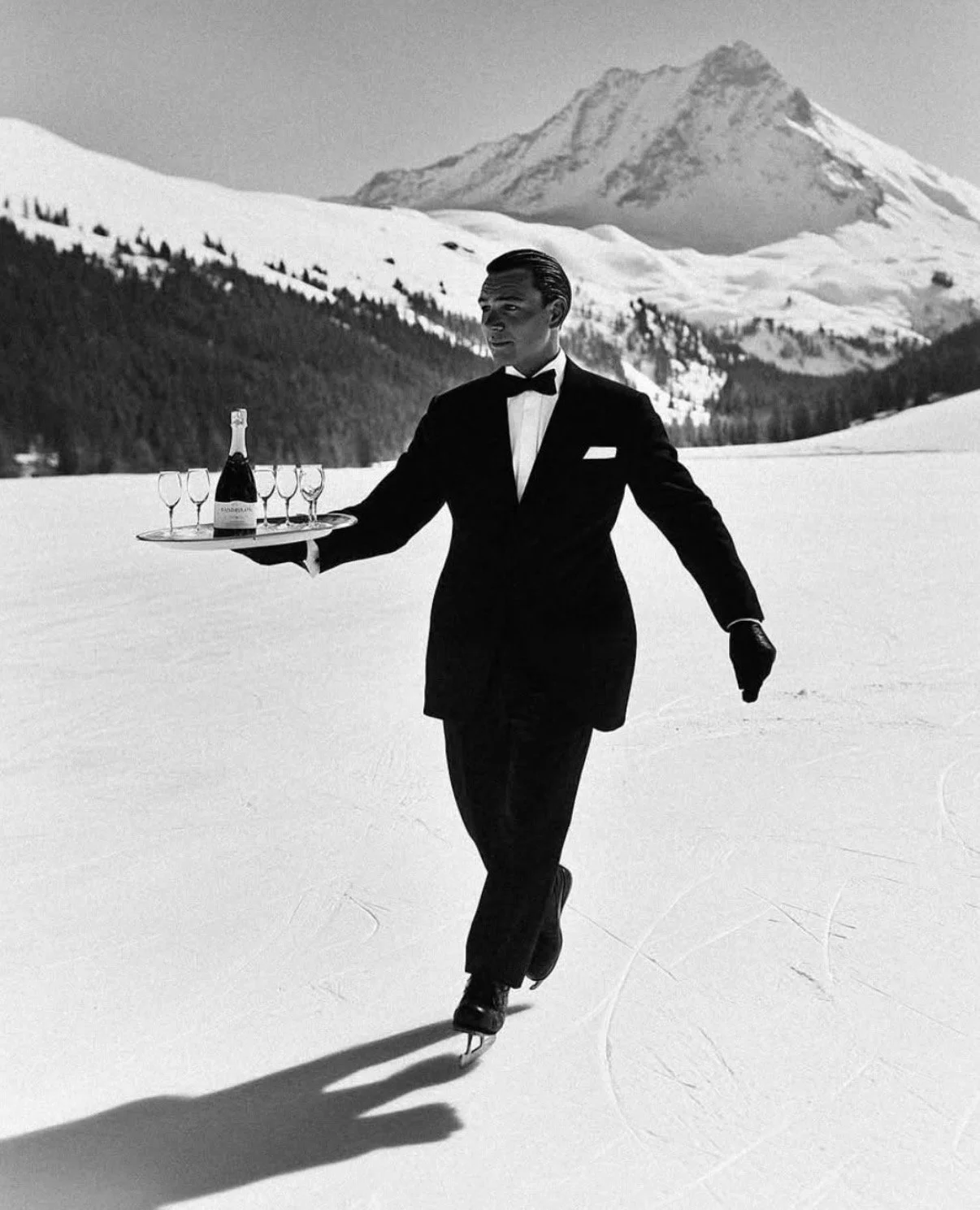 A man in a tuxedo ice skating on snow-covered ground, holding a tray with a bottle of champagne and six champagne glasses, with a mountain and forest in the background.