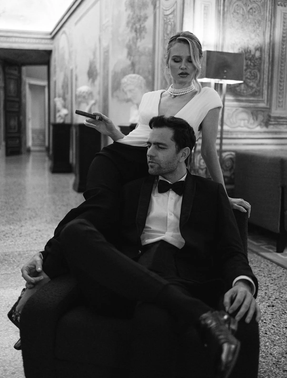 A woman in an elegant dress and pearl necklace is standing behind a seated man in a tuxedo, in an ornate, classical interior.