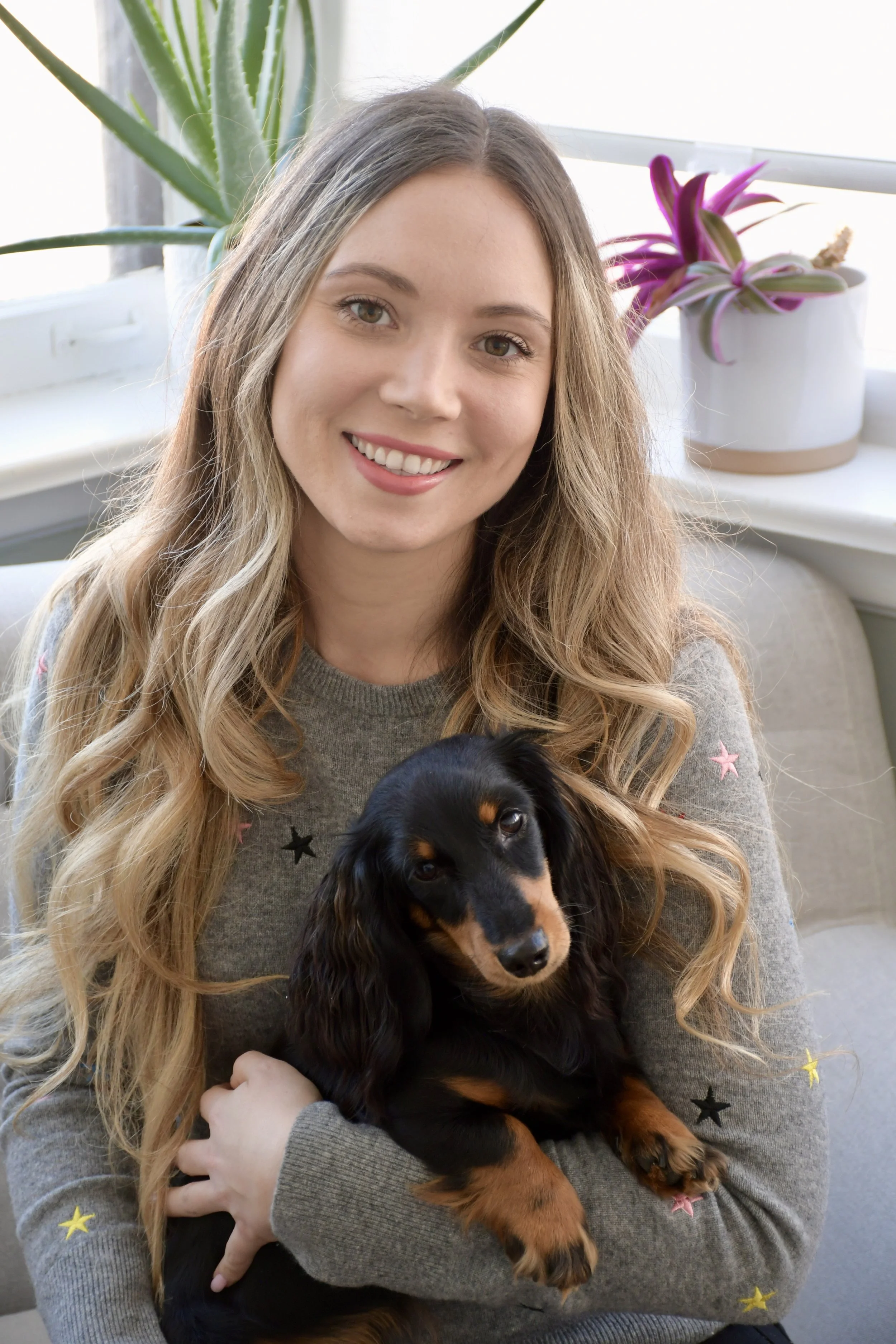 A young woman with long wavy blonde hair smiling while holding a black and tan dachshund puppy indoors in front of a white windowsill with potted plants.