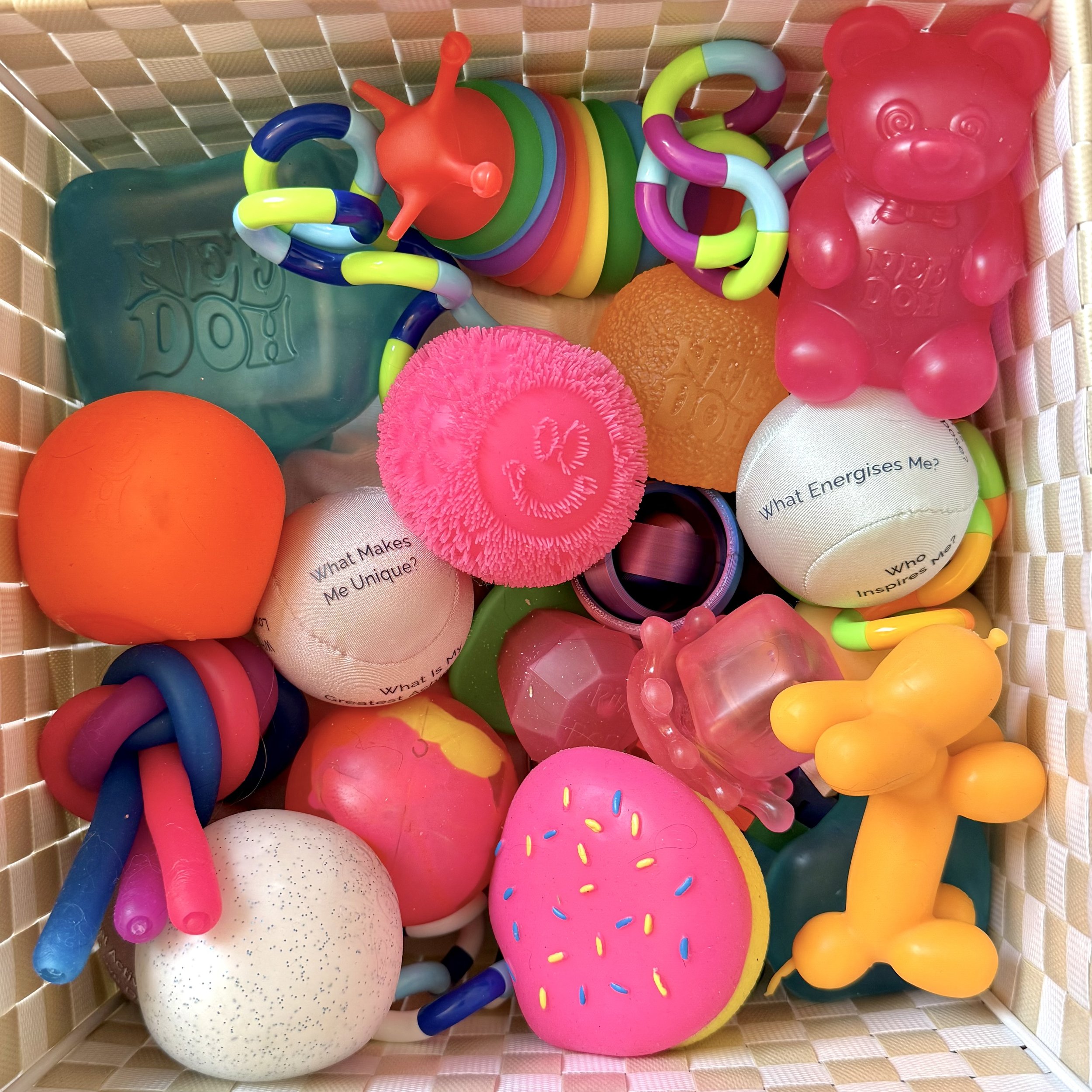 A collection of colorful children's toys including a pink foam donut, a pink bear-shaped soap, a yellow balloon animal, a plush ball with a smiley face, a large orange foam sphere, a turquoise soap with embossed text, various textured balls, Winnie the Pooh-shaped soap, and rainbow-colored rings.