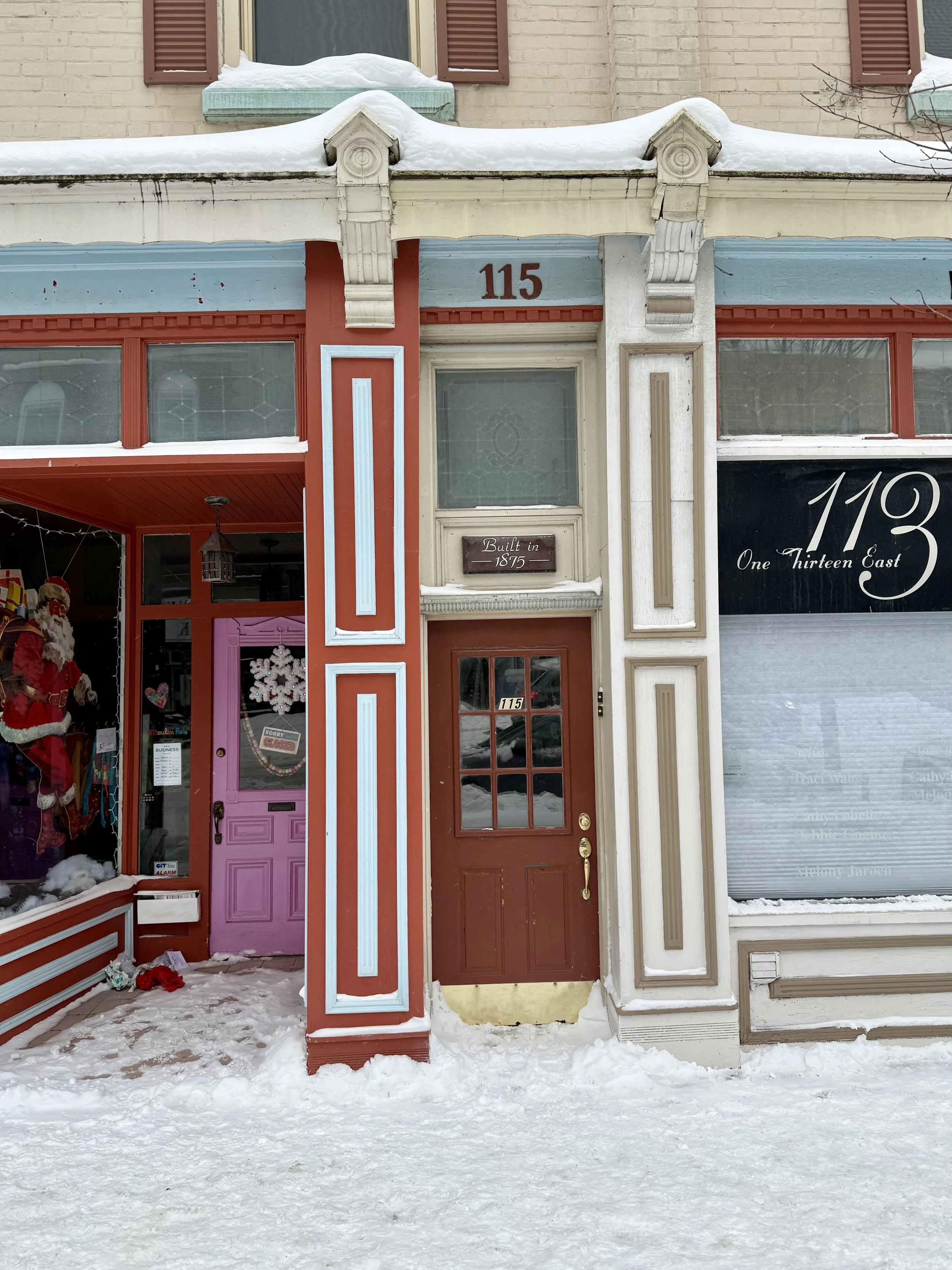The storefront of 115 One Thirteen East with a red door, decorative white and blue trim, and snow on the ground.