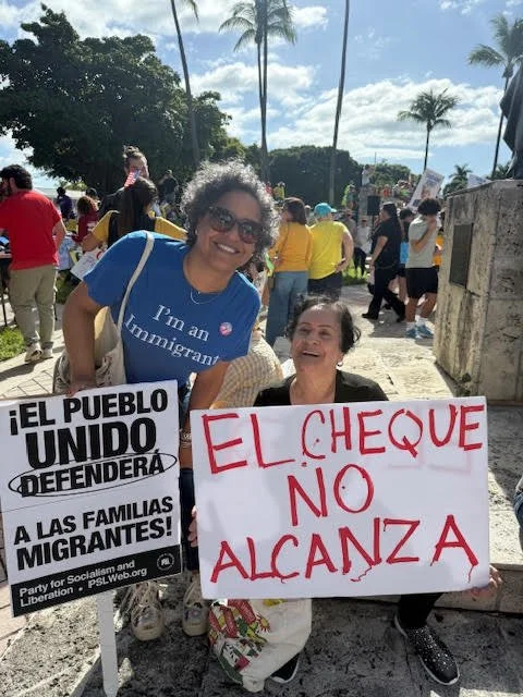 Two women participate in a demonstration with signs protesting migration issues, surrounded by other protesters in a tropical outdoor setting with palm trees.