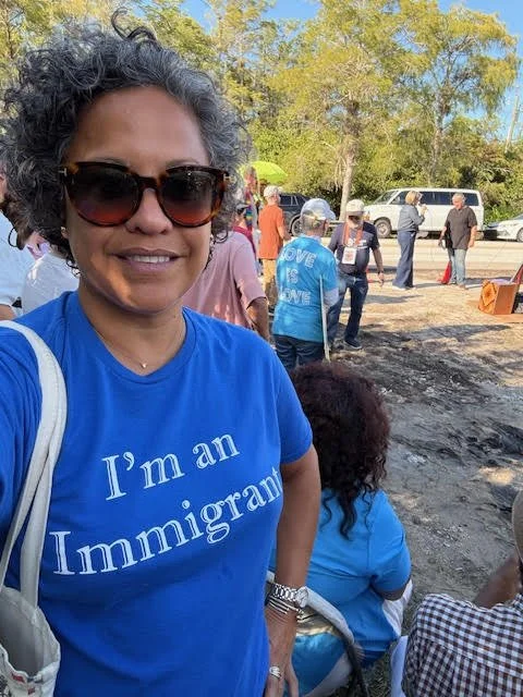 Yurina Gil wearing sunglasses and a blue t-shirt that says 'I'm an Immigrant,' standing outdoors with a group of people and cars in the background.