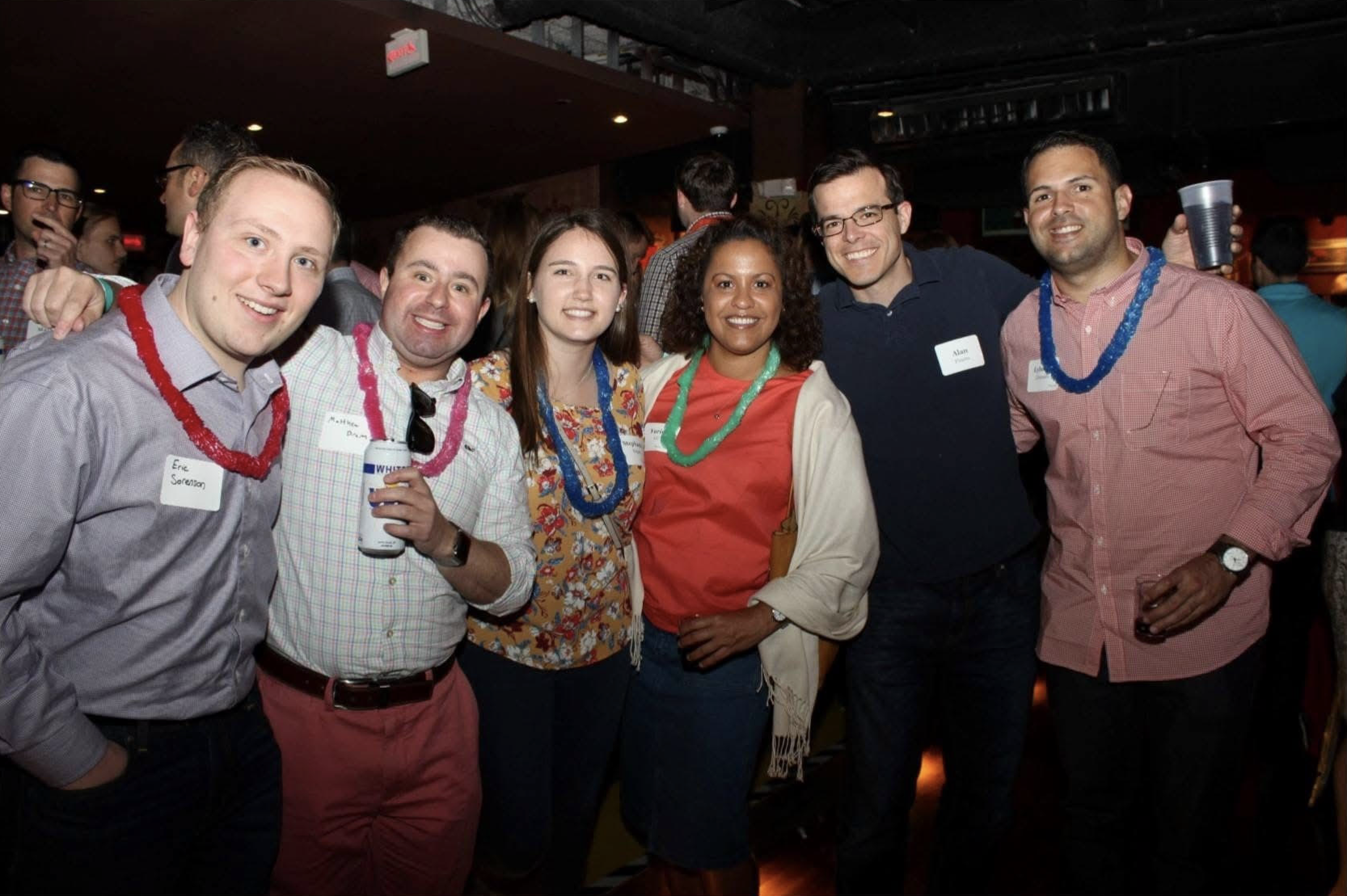 Group of seven people at party wearing colorful leis and smiling, with some holding drinks.