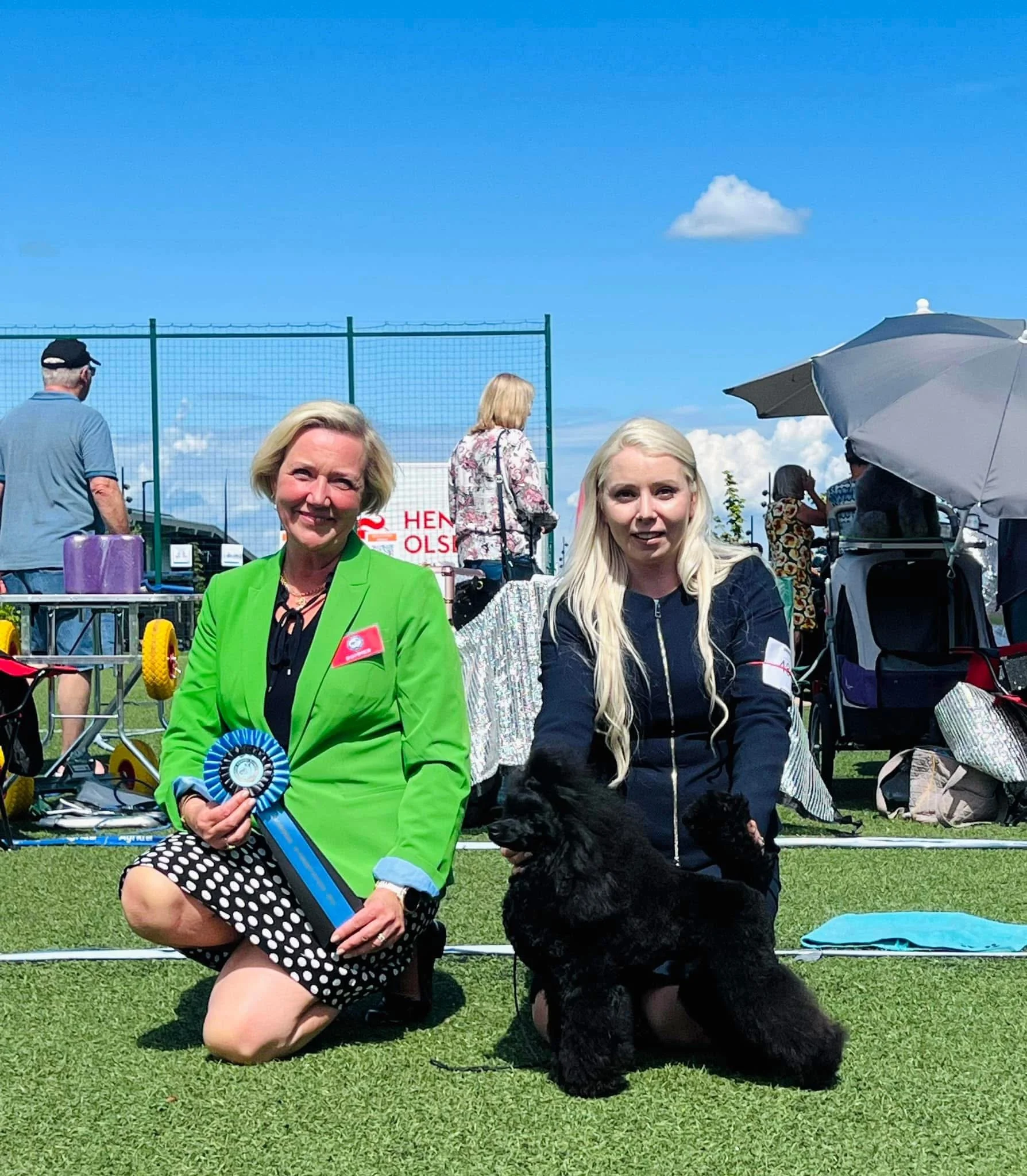 Two women, one in a bright green blazer and polka-dot skirt, and the other in a black jacket, kneeling on grass at an outdoor event with a black dog, possibly a poodle, between them. The woman in green is holding a blue and white ribbon. People and t