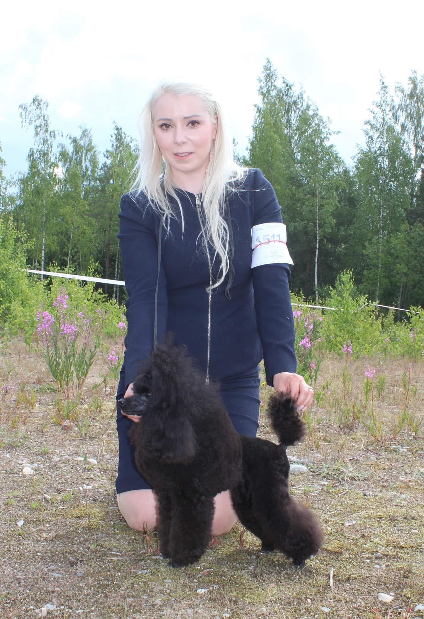 A woman with long blonde hair kneeling outdoors in a field, holding a black poodle, with trees and pink flowers in the background.