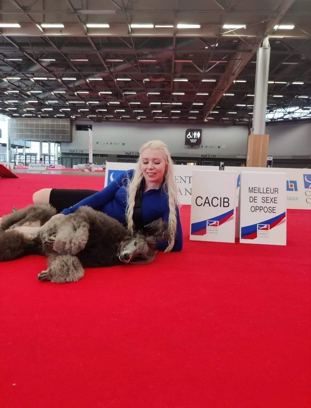 A woman lying on a red carpet with a relaxed dog, possibly a wolfdog, beside her, at a dog competition or event in a large indoor arena with signs displaying French text and a ceiling with metal beams and lighting.