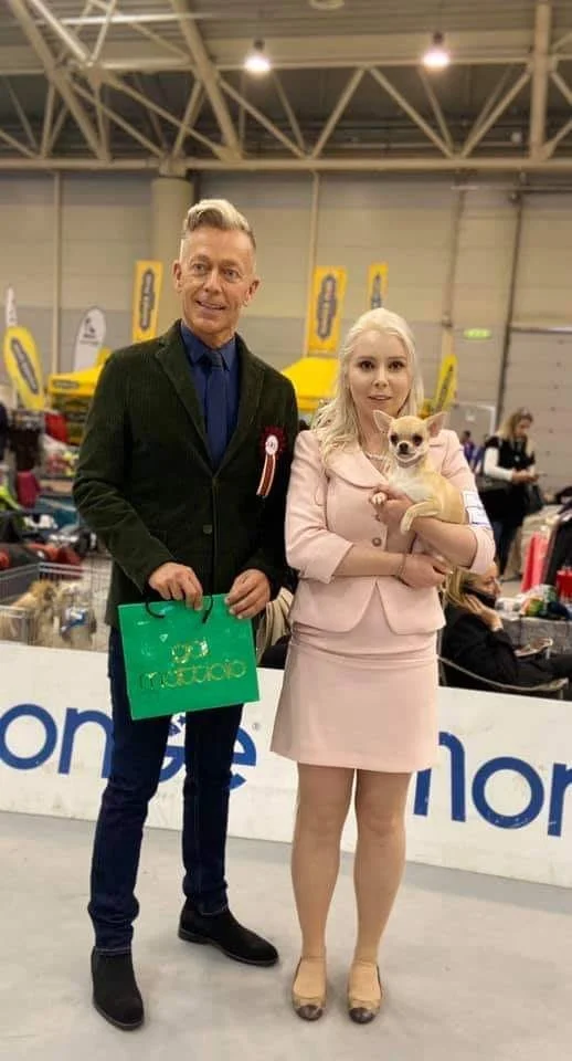 A man and a woman with a Chihuahua dog at an indoor event or exhibition.