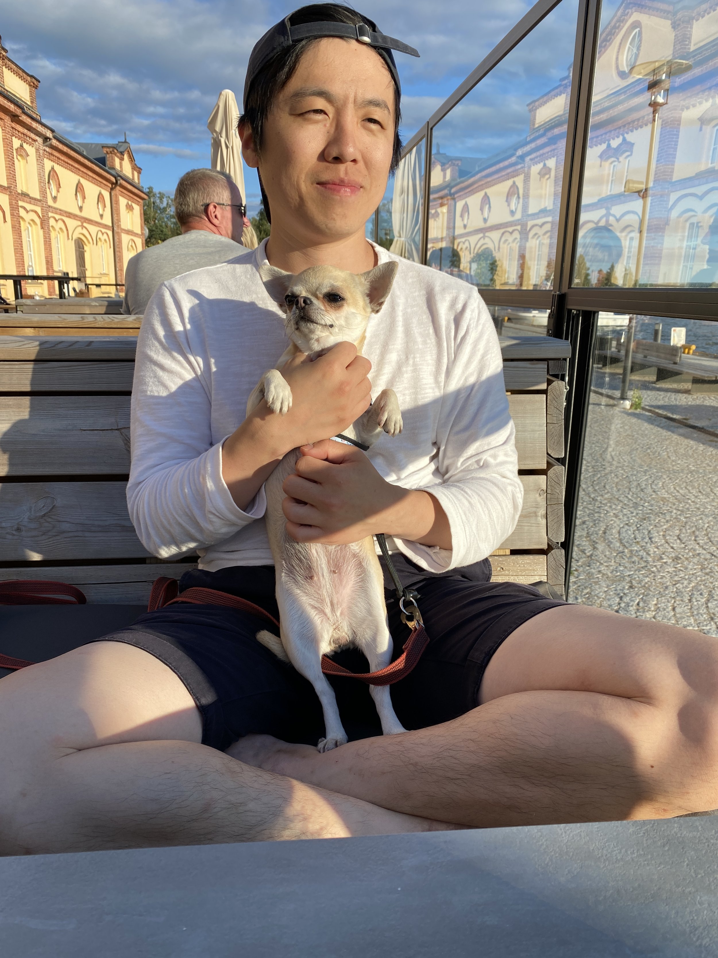 A young man sitting outdoors on a wooden bench, holding a small dog with a light tan coat, against a background of historic European-style buildings and a glass barrier, during late afternoon sunlight.