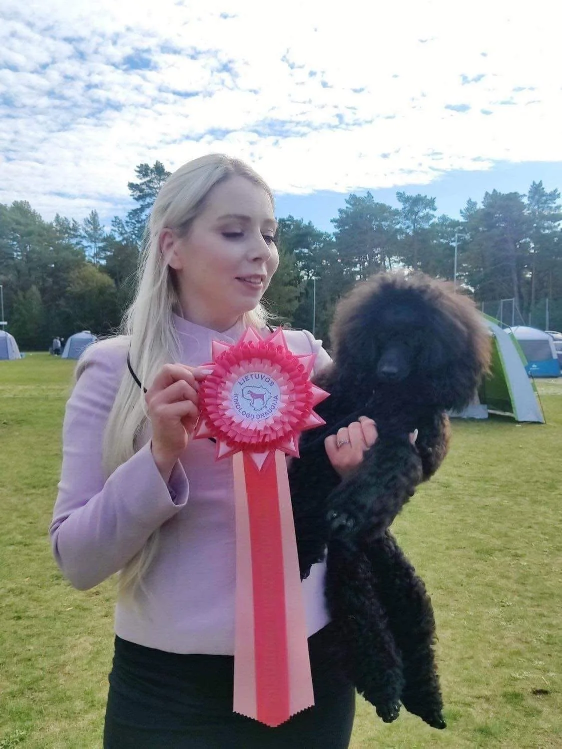 A woman holding a black poodle and a pink ribbon award outdoors on a grassy field with trees and tents in the background.