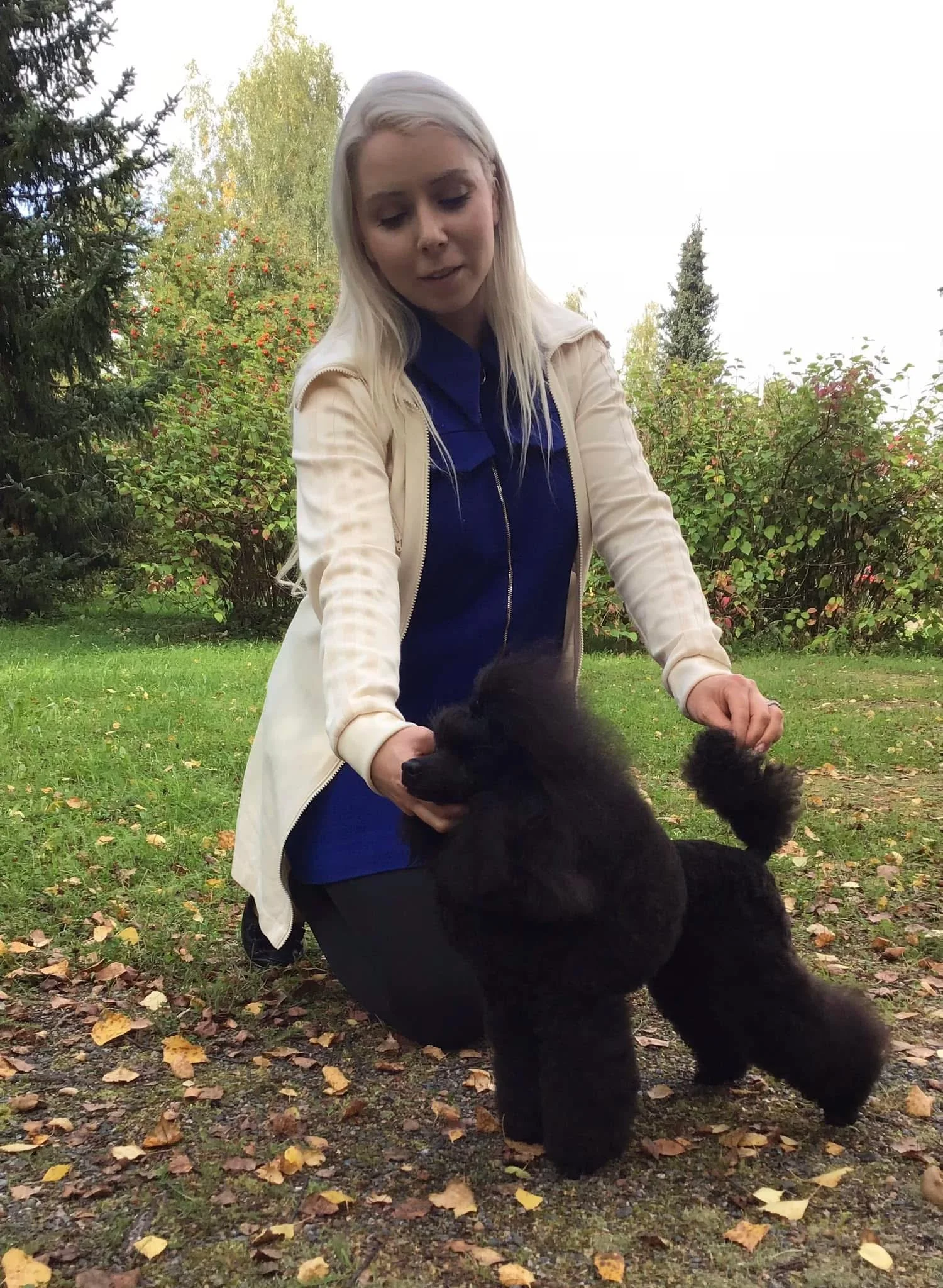 A woman with long blonde hair wearing a cream-colored jacket and a dark blue shirt, kneeling on grass with fallen leaves, holding a black fluffy poodle outdoors in a park with trees in the background.