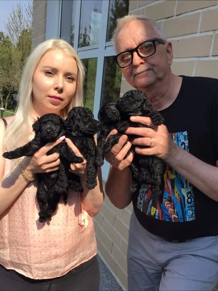A young woman and an older man are holding black puppies outside a building with large windows and a brick wall.