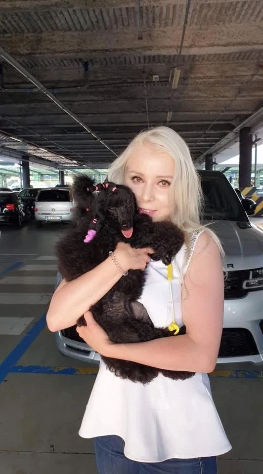 A woman with long blonde hair holding a black poodle puppy with pink accessories in a parking garage.