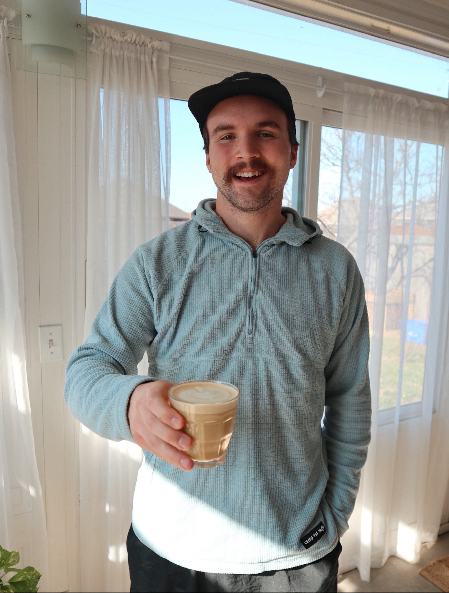 A man smiling and holding a glass of coffee. He is wearing a gray hoodie, a black cap, and standing inside near windows with white curtains.