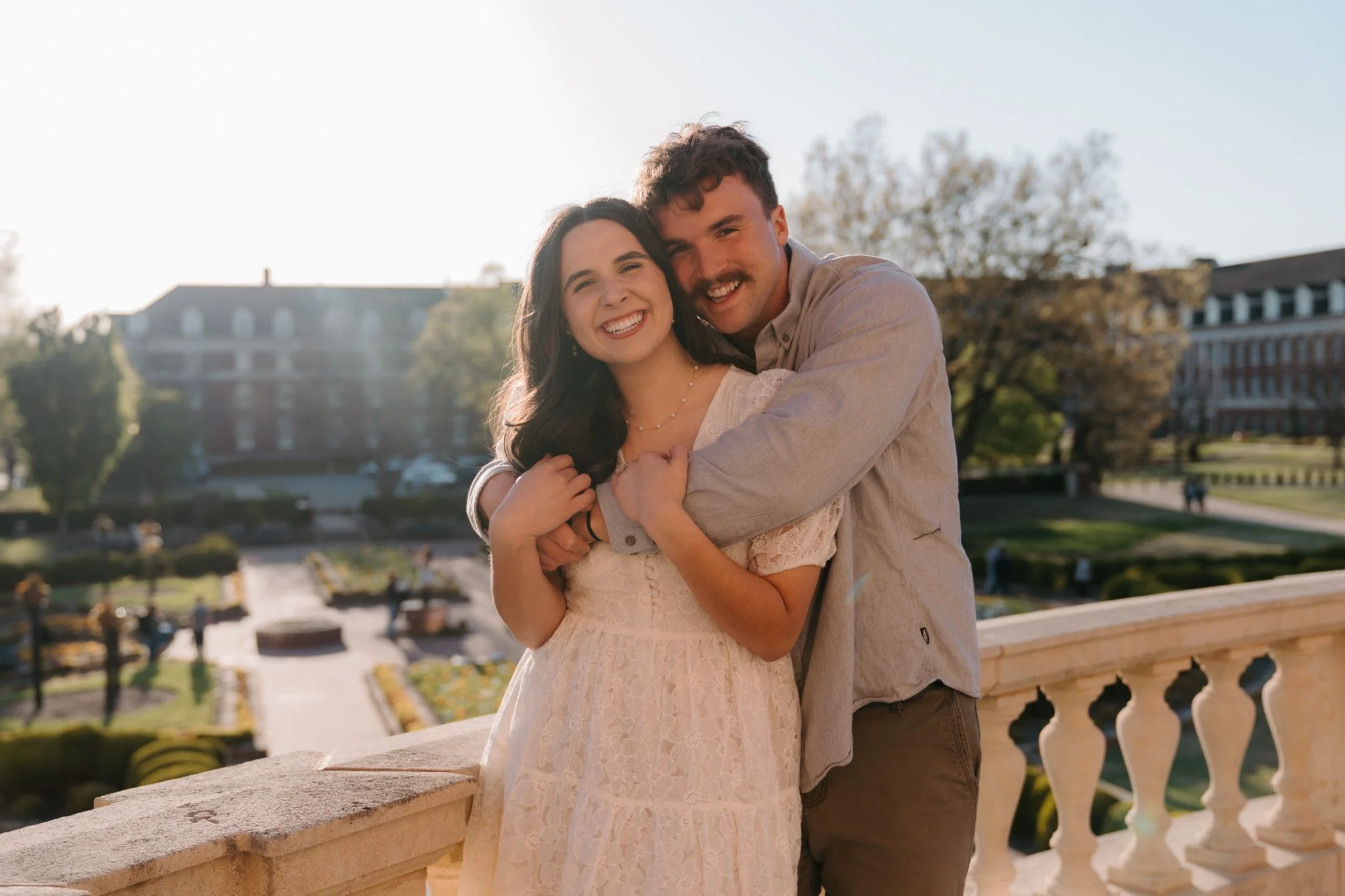 A smiling young couple hugging on a balcony overlooking a park or garden during sunset, with buildings and trees in the background.