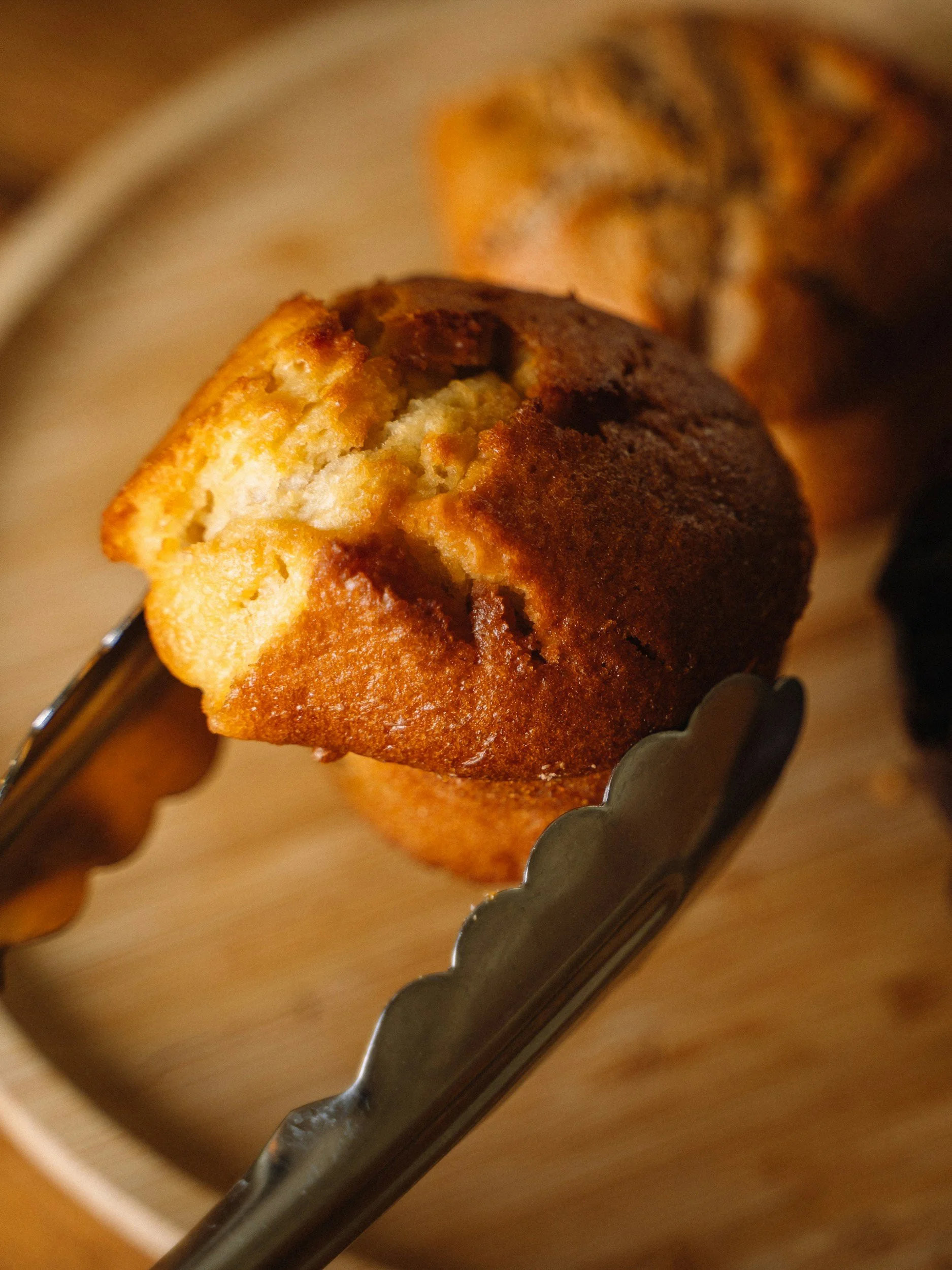 A close-up of a freshly baked vegan muffin held with metal tongs, with another muffin in the background, on a wooden surface.