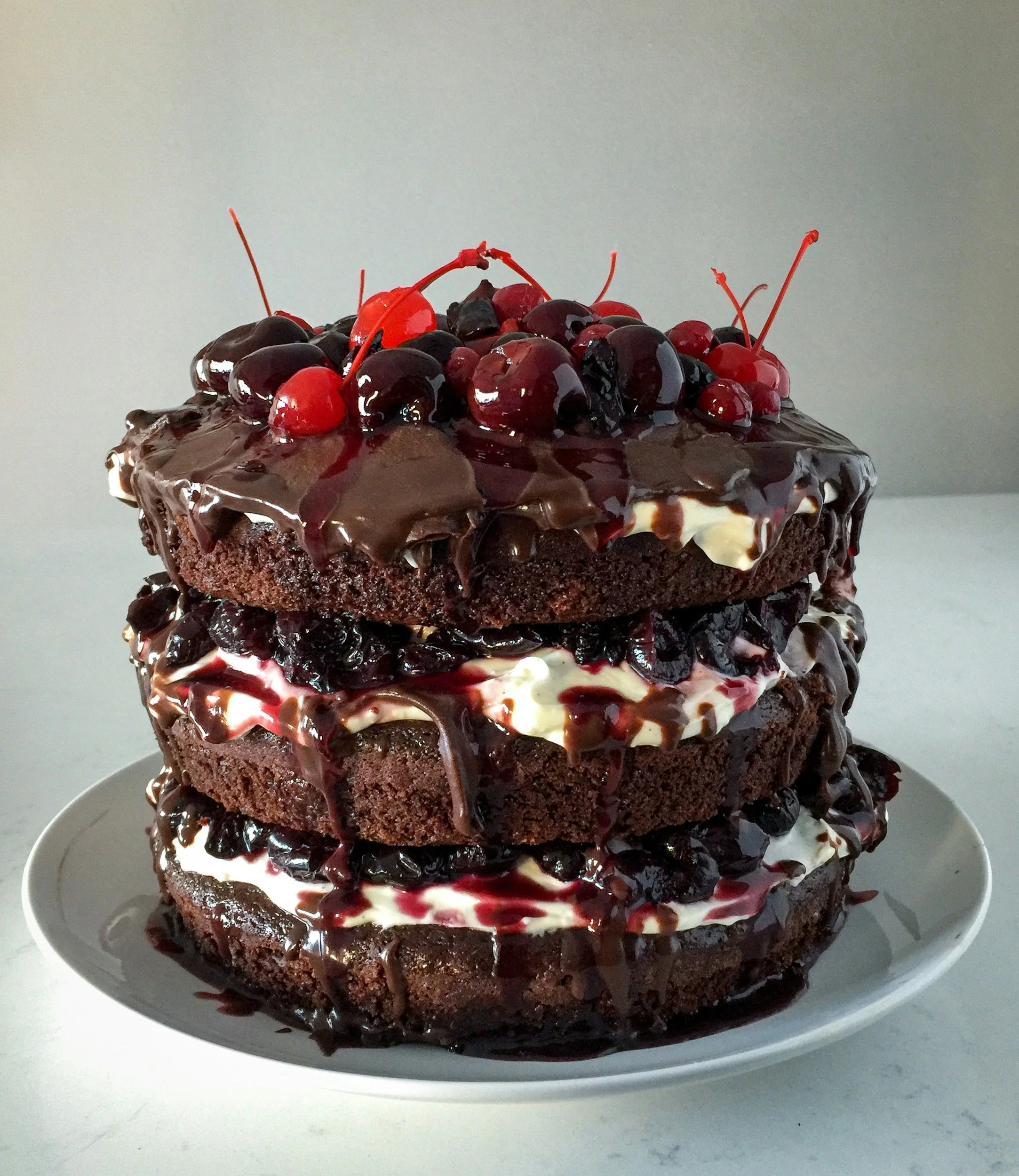 Three-layer vegan chocolate cake with chocolate and berry toppings, drizzled with chocolate sauce, on a white plate.