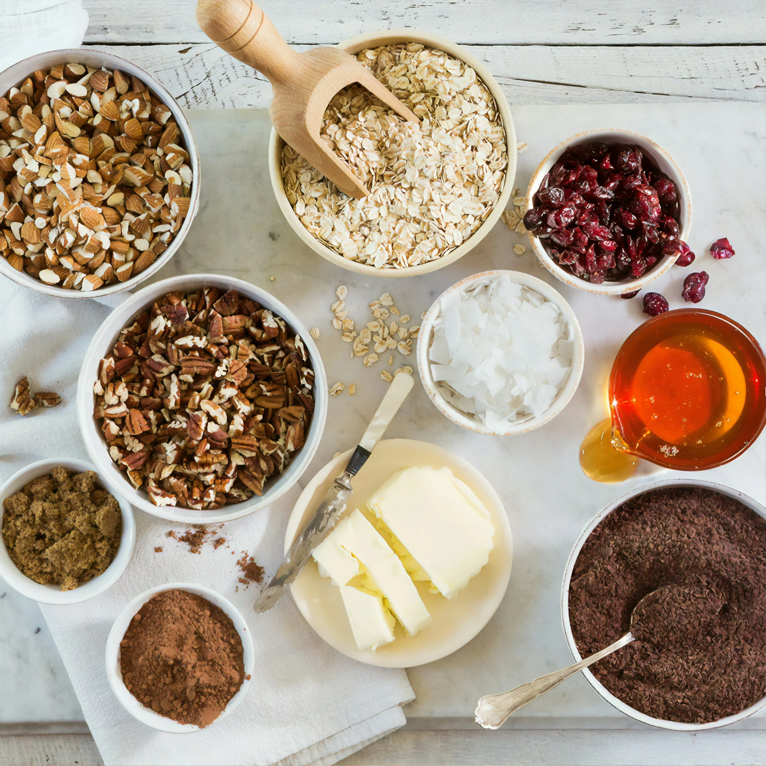 Various bowls of ingredients for baking, including chopped nuts, oats, dried cranberries, shredded coconut, chocolate chips, cocoa powder, white butter, brown sugar, and liquid honey, arranged on a marble surface.