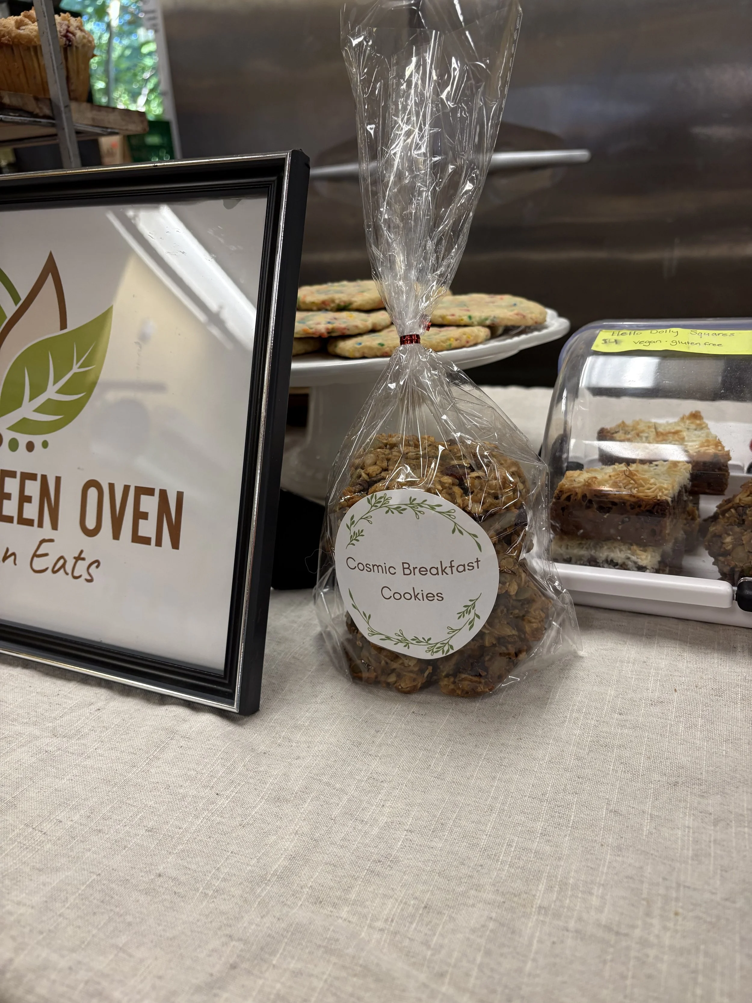 A clear plastic bag of granola labeled 'Cosmic Breakfast Cookies' on a table at an event. In the background, there are trays of cookies, a sign that reads 'Green Oven, Vegan Eats,' and various baked goods.