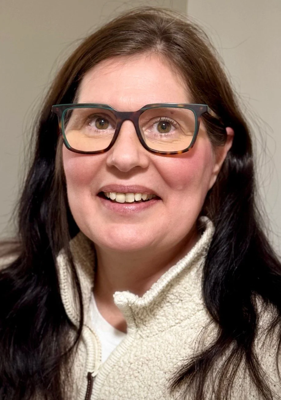 A woman with long dark hair, wearing glasses and a cream-colored fleece jacket, smiling at the camera.