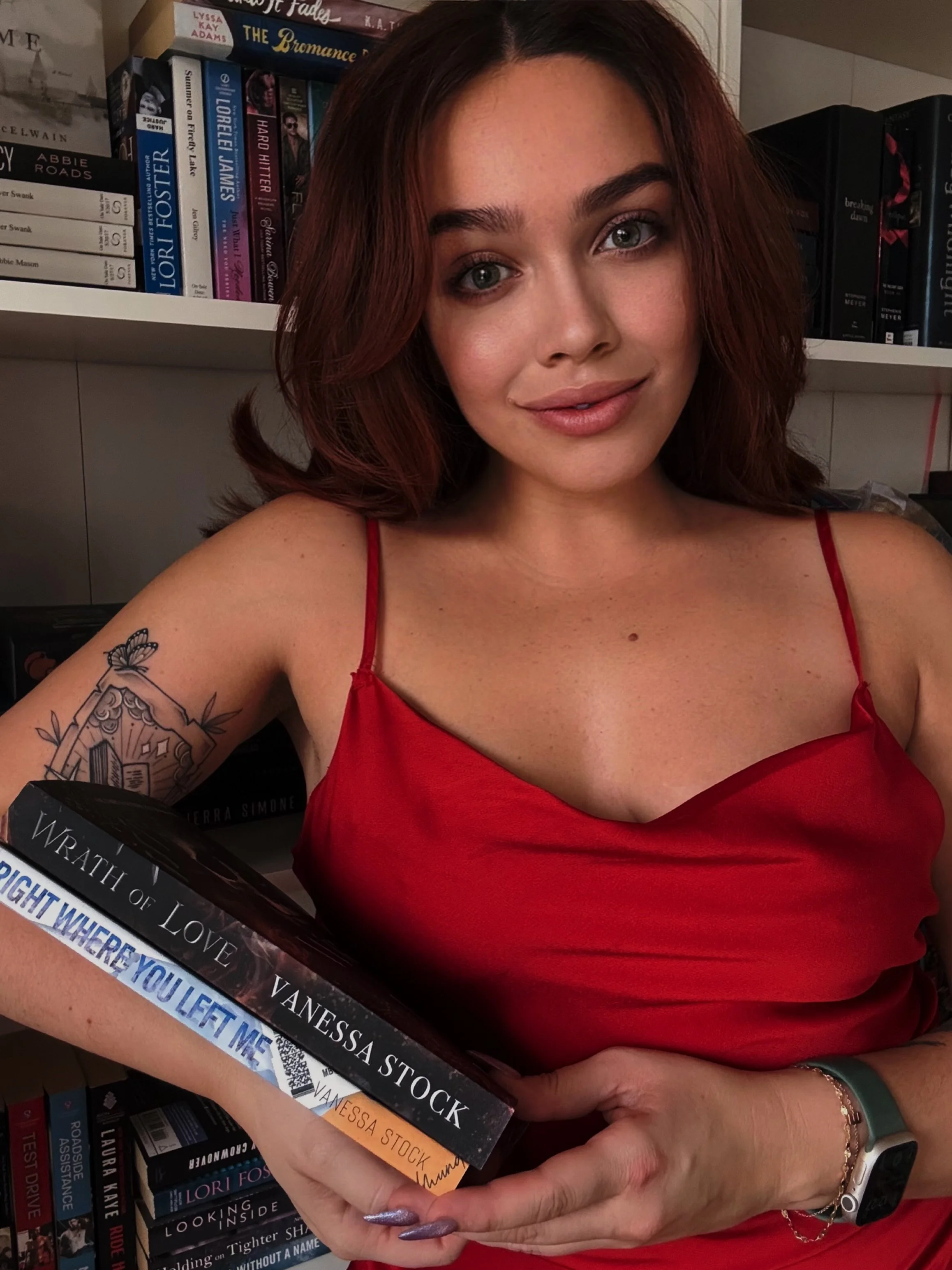 A young woman with shoulder-length red hair and green eyes, wearing a red spaghetti strap top, holding a book titled "Right Where You Left Me" by Vanessa Stock, and standing in front of bookshelves filled with various books.