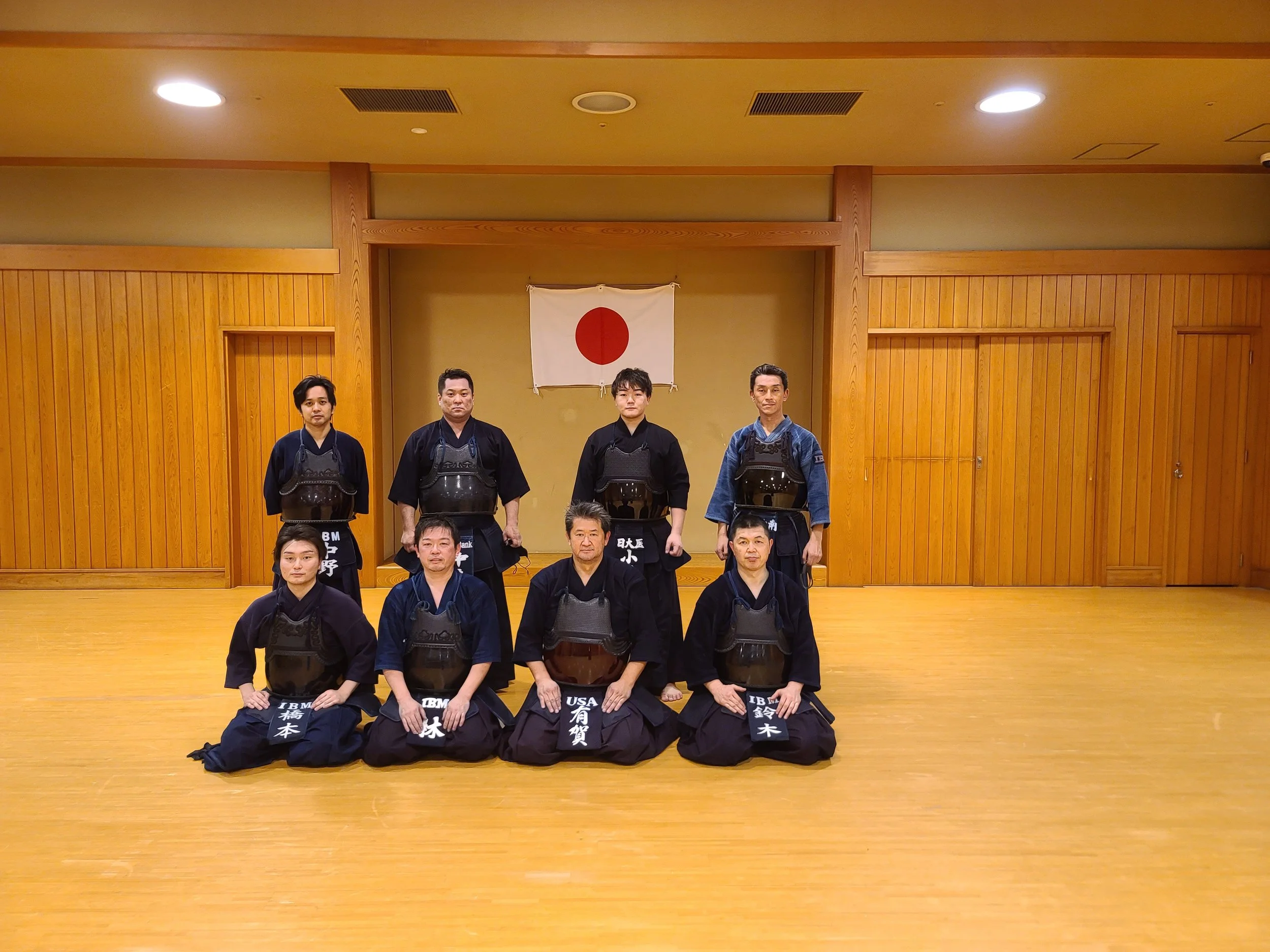 Group of eight martial artists wearing traditional kendo uniforms and armor, kneeling and standing in a dojo with wood paneling and a Japanese flag behind them.