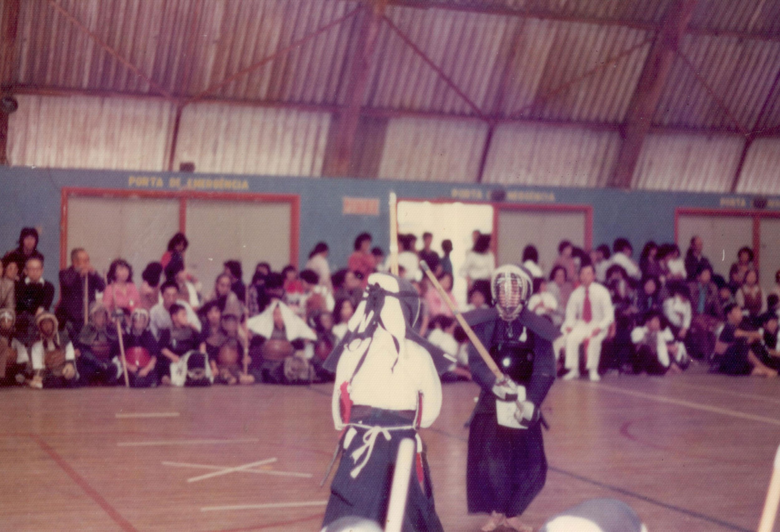 A traditional martial arts demonstration in a gymnasium with an audience watching; two practitioners wearing black uniforms and protective gear engage in a sparring match, with one holding a wooden sword and the other wielding a staff, surrounded by spectators in the background.