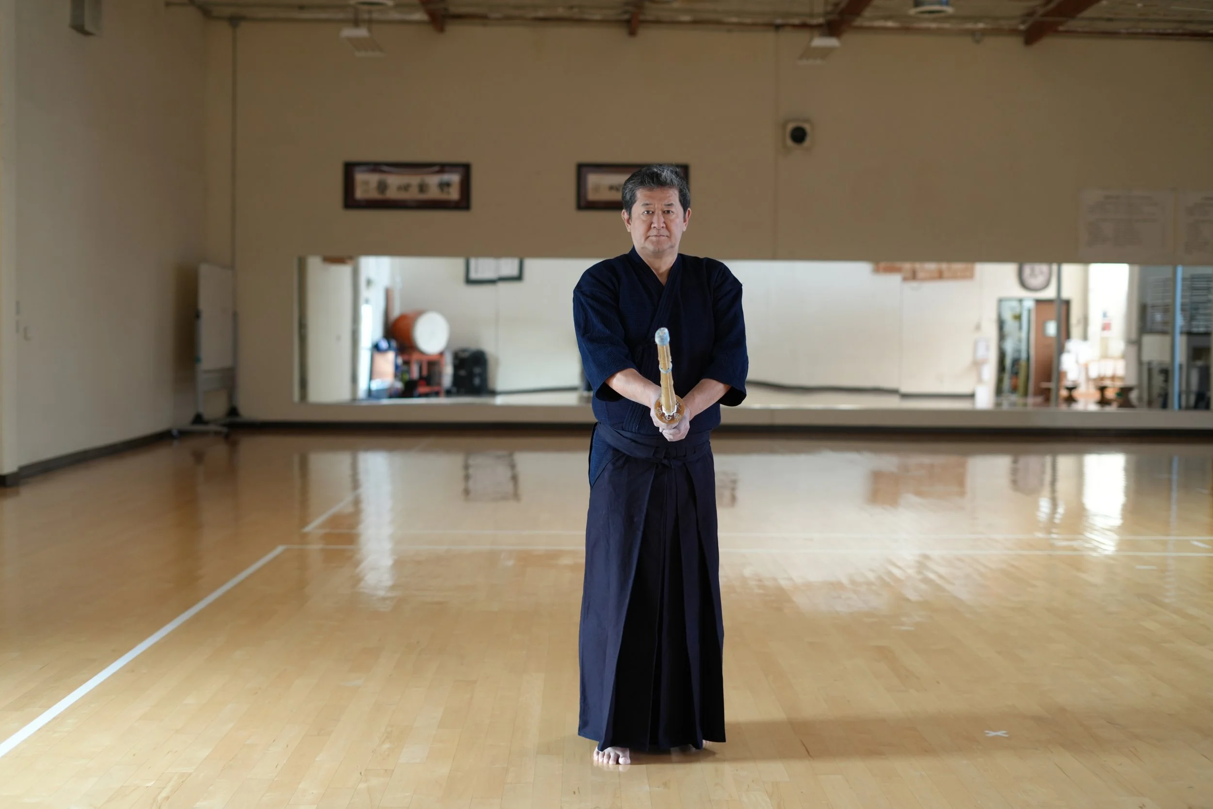 A man dressed in traditional martial arts attire holding a wooden sword in an empty dojo with a mirror on the back wall.