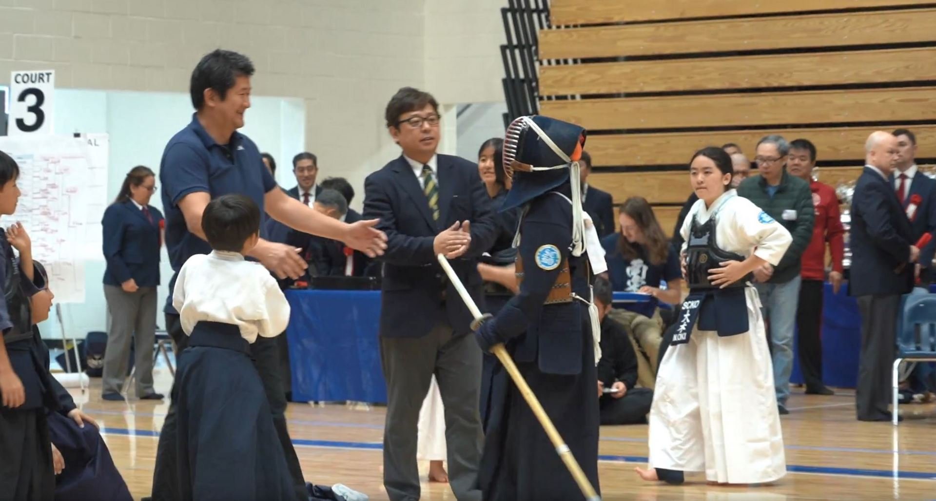 People in a gymnasium, some wearing martial arts uniforms, shaking hands and interacting during a martial arts event or competition.