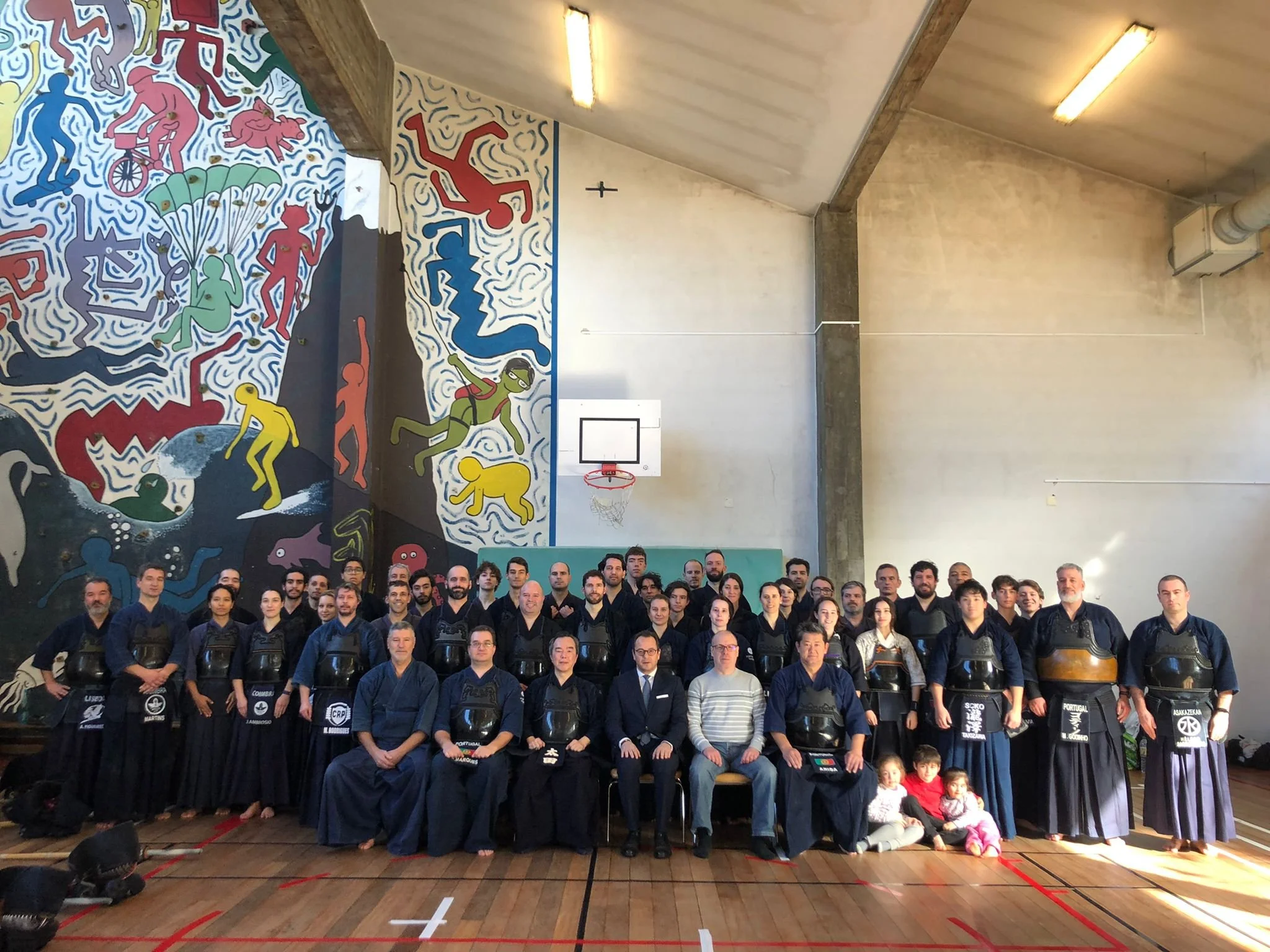 A group of people in traditional martial arts uniforms posing in a gymnasium with a colorful mural on the wall behind them.