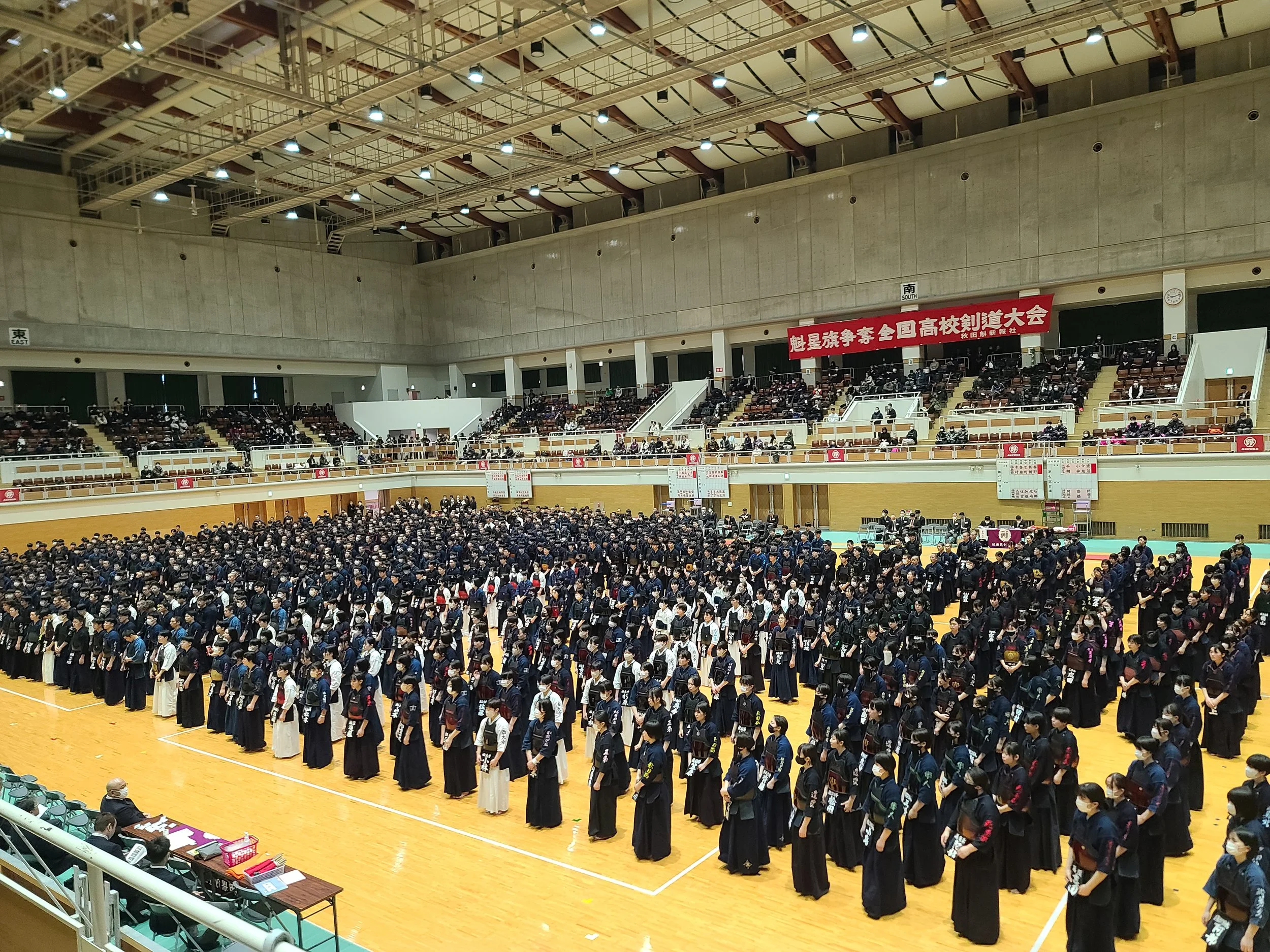 Hundreds of people in traditional kendo uniforms standing in formation inside a gymnasium during a large event, with spectators seated around the perimeter and a red banner with Japanese characters hanging on the wall.