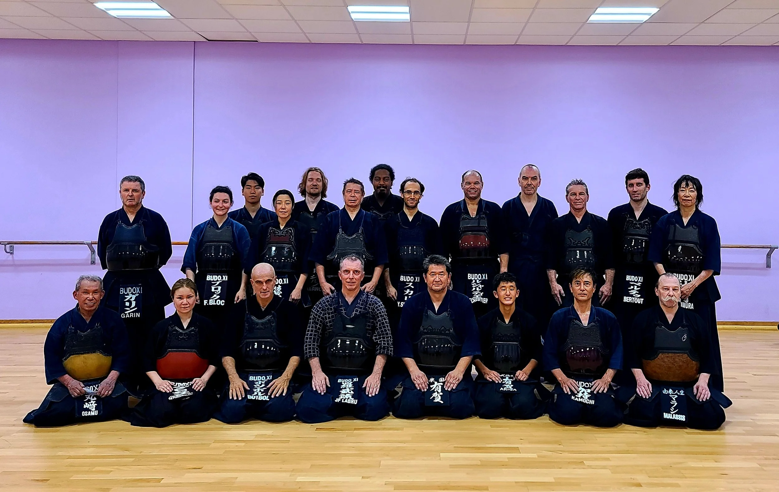 Group photo of martial artists in traditional uniforms, sitting and standing in a dojo with purple walls and wooden floors.