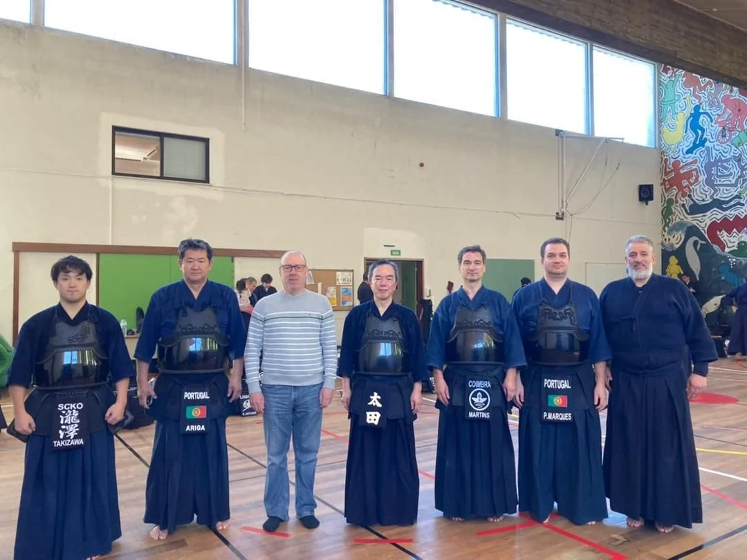 Group of seven men in martial arts uniforms standing in a gymnasium, some with striped clothing, with a colorful mural on the wall in the background.