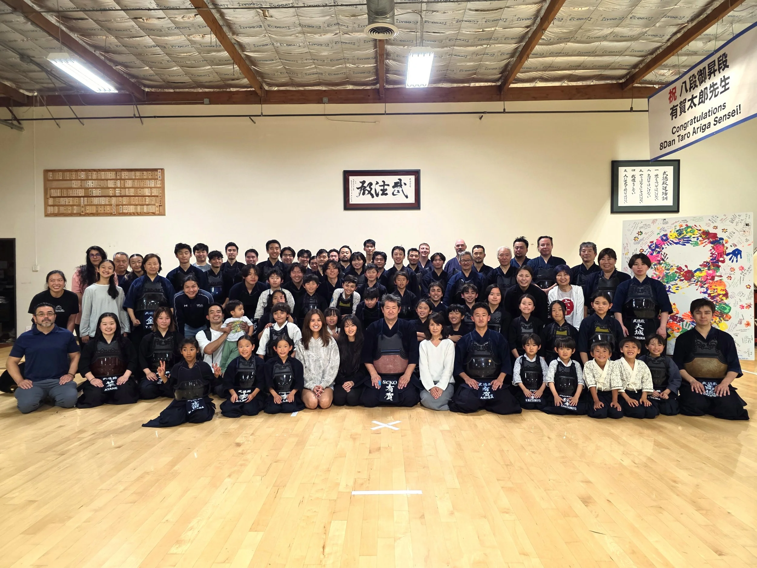Group of people, including children and adults, posing for a photo inside a martial arts dojo, with some wearing traditional kendo gear and armor, and banners with Japanese writing on the walls.