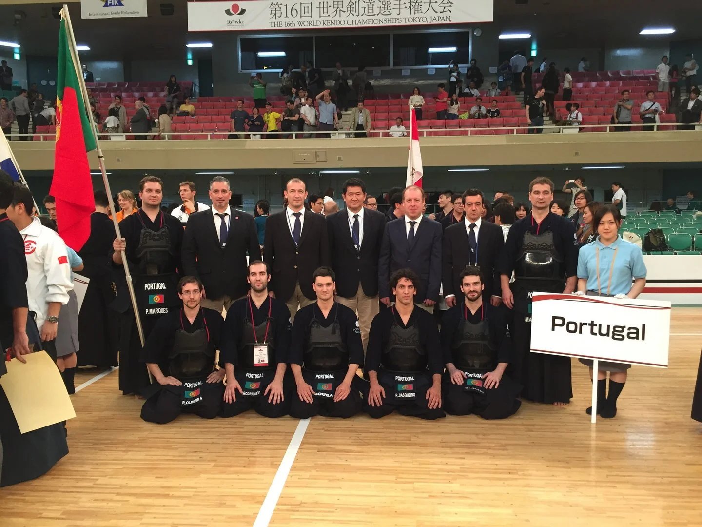A group of athletes and officials from Portugal at the 16th World Kendo Championships in Tokyo, Japan. They are dressed in traditional kendo uniforms and are posing for a photo with some holding the Portuguese flag. The scene takes place inside an indoor sports arena with other participants and spectators in the background.