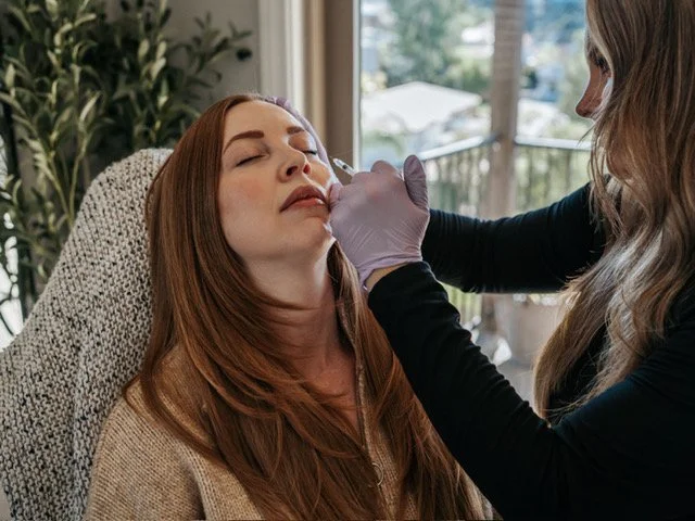 A woman receives a cosmetic treatment, possibly a Botox injection, from a professional wearing black gloves in a well-lit room with large windows and greenery outside.