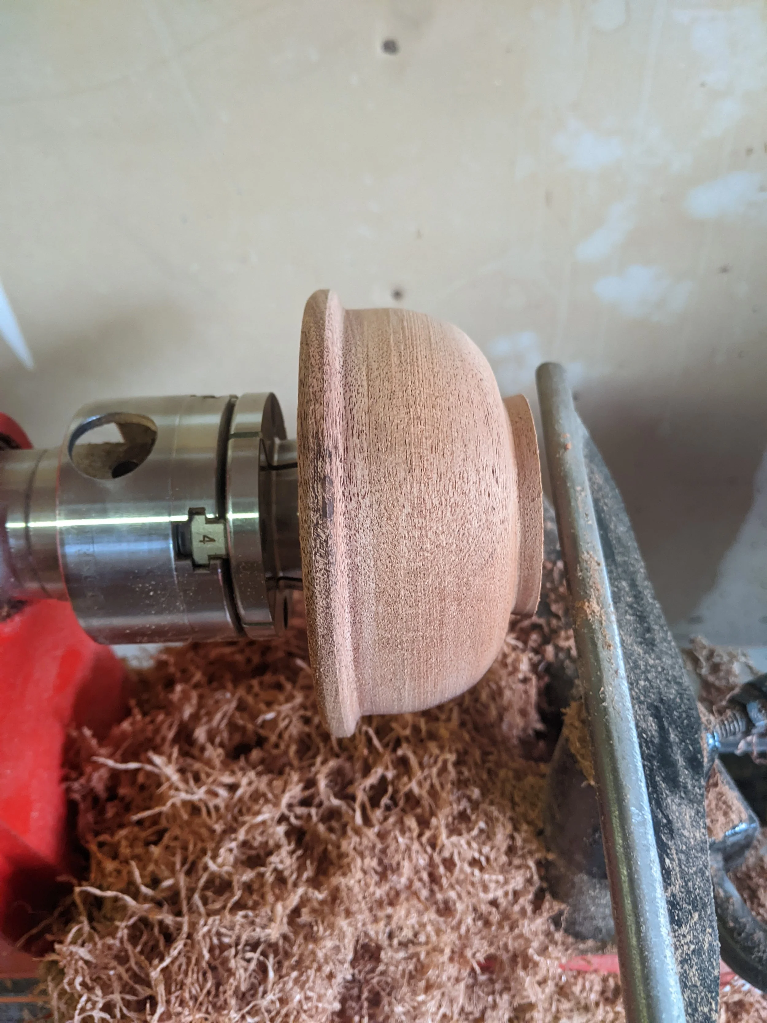 Close-up of a woodturning project on a lathe, showing a partially shaped wooden piece with a rounded top and a flat bottom, surrounded by wood shavings.
