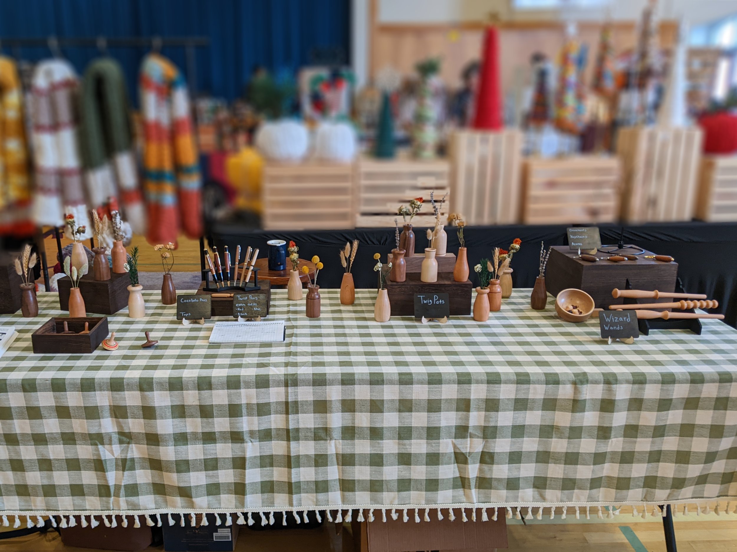 A local craft fair display table filled with wood home decor and art. Main items include small wood vases, pens, and decorated spinning tops.