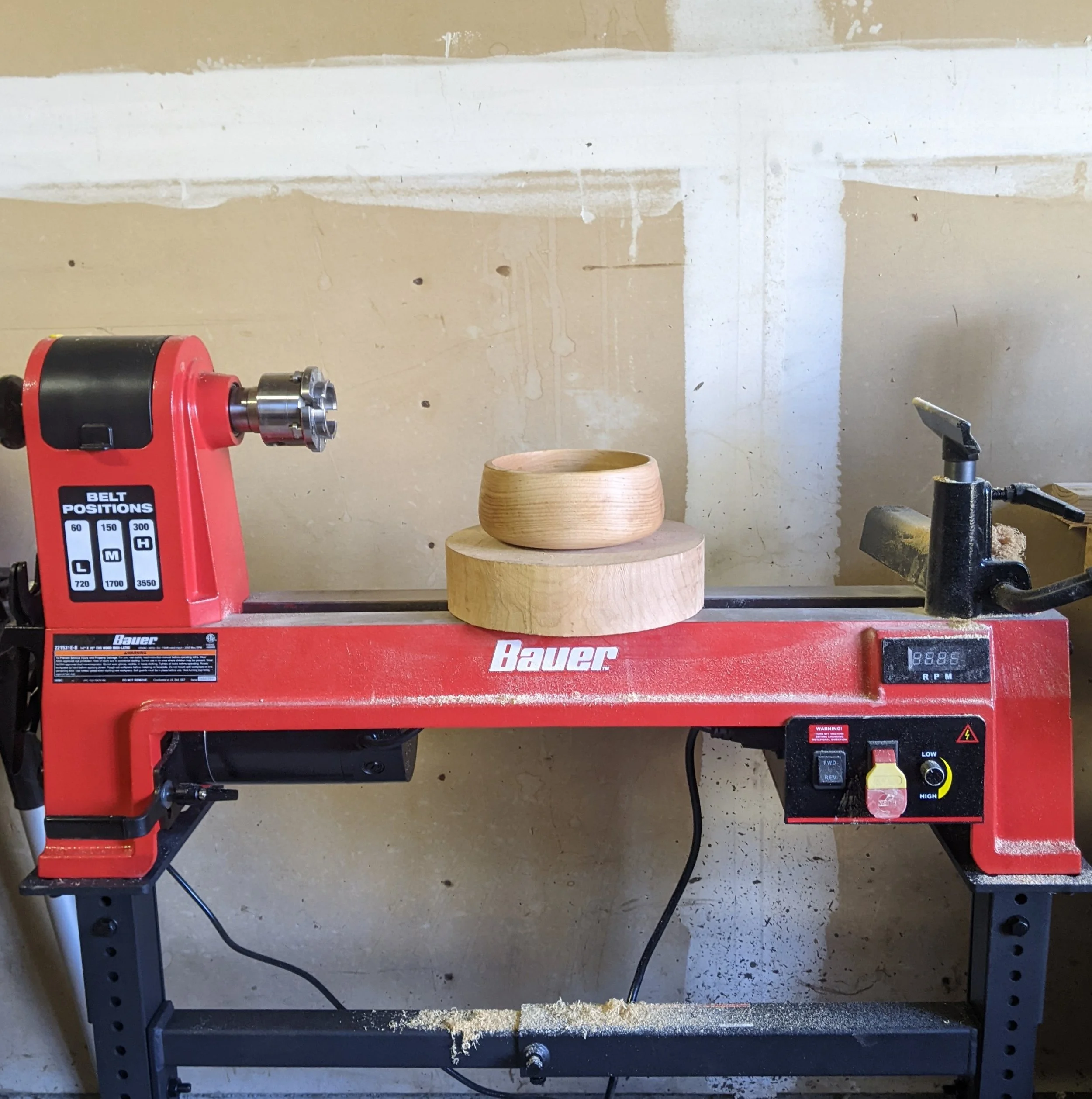 Red woodworking lathe with a finished wooden bowl and a bowl blank on top, set against a beige wall.