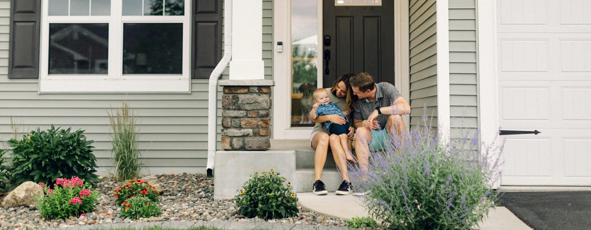 A family sitting on front porch steps, smiling and playing with a young child; house with gray siding, black shutters, and a white garage door in the background.