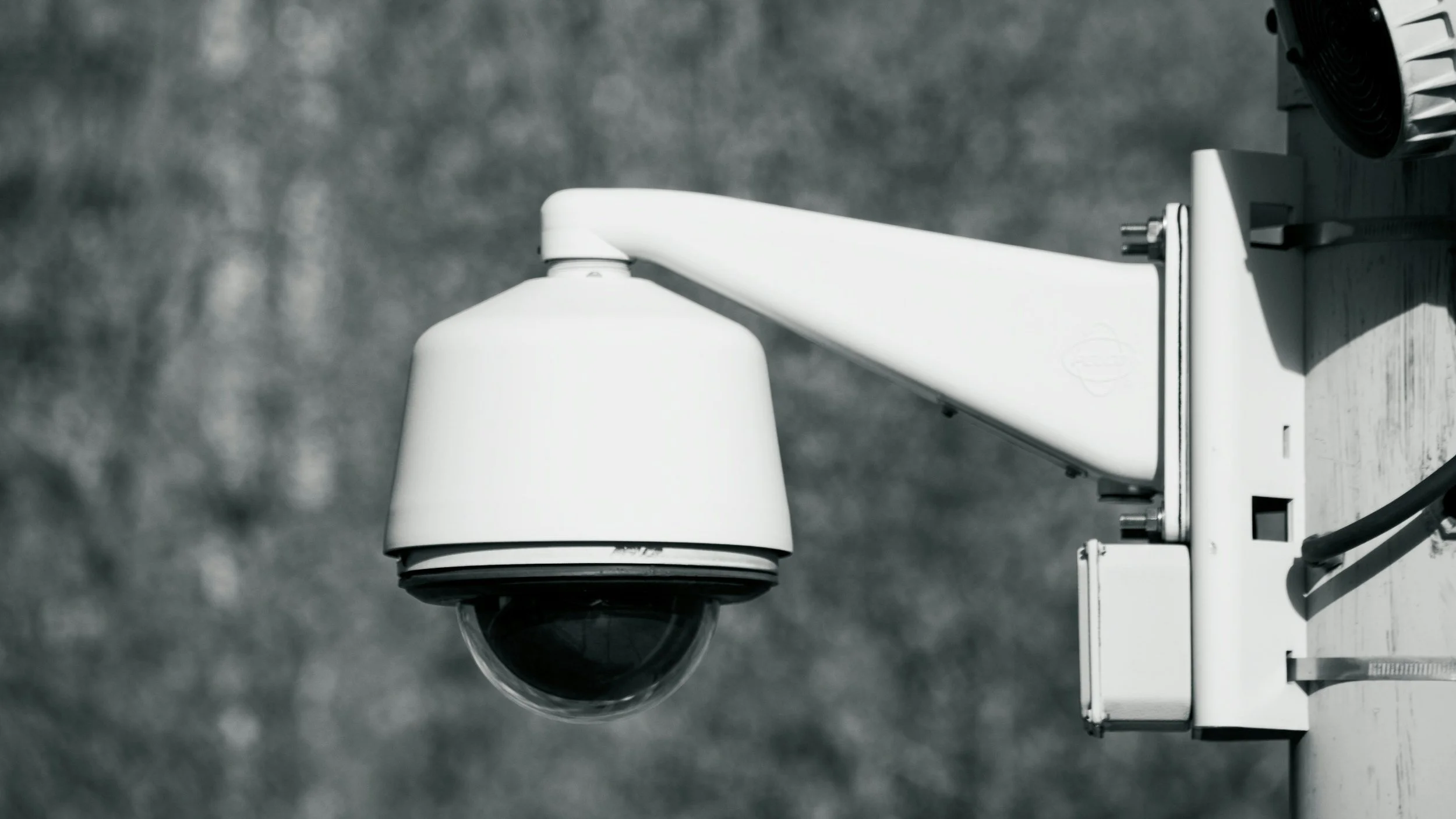 Security camera mounted on a building, pointing downward, with a blurred outdoor background.