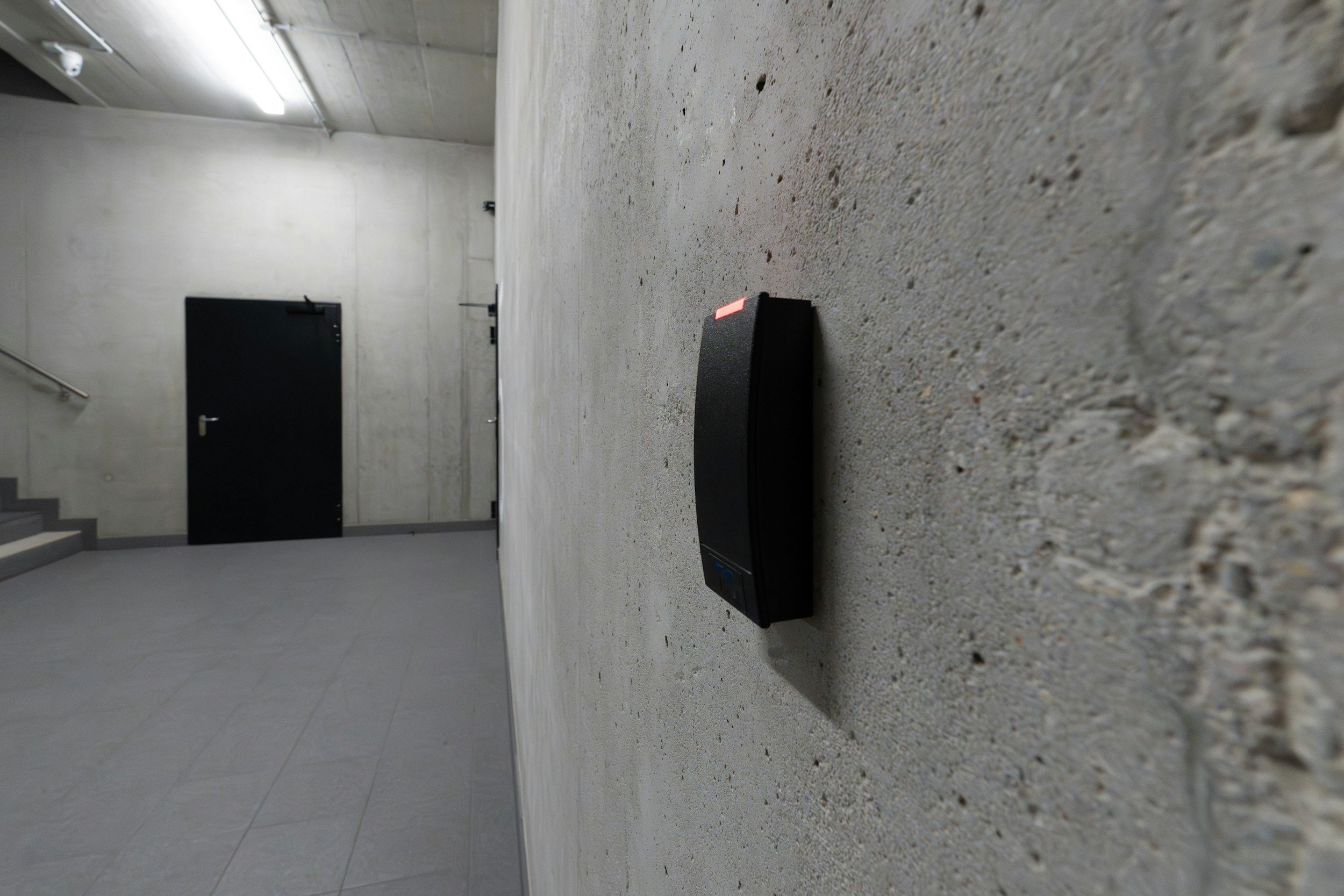 A black rectangular device is placed on a concrete floor in a gray, industrial-style hallway with stairs and a black door in the background.